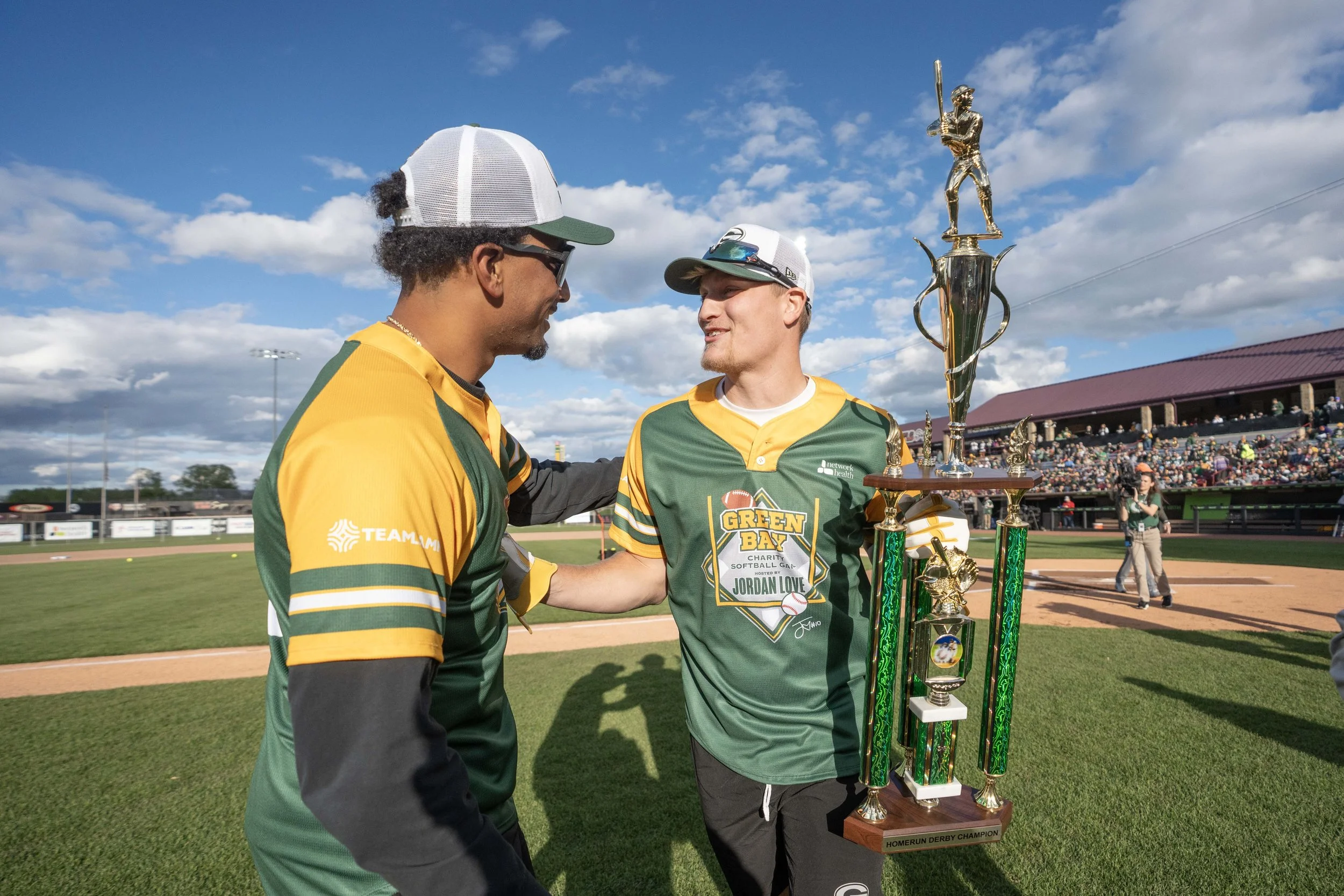 Two young men in green and yellow baseball jerseys shaking hands on a baseball field, one of them holding a large trophy, with a crowd in the stands and a partly cloudy sky overhead.