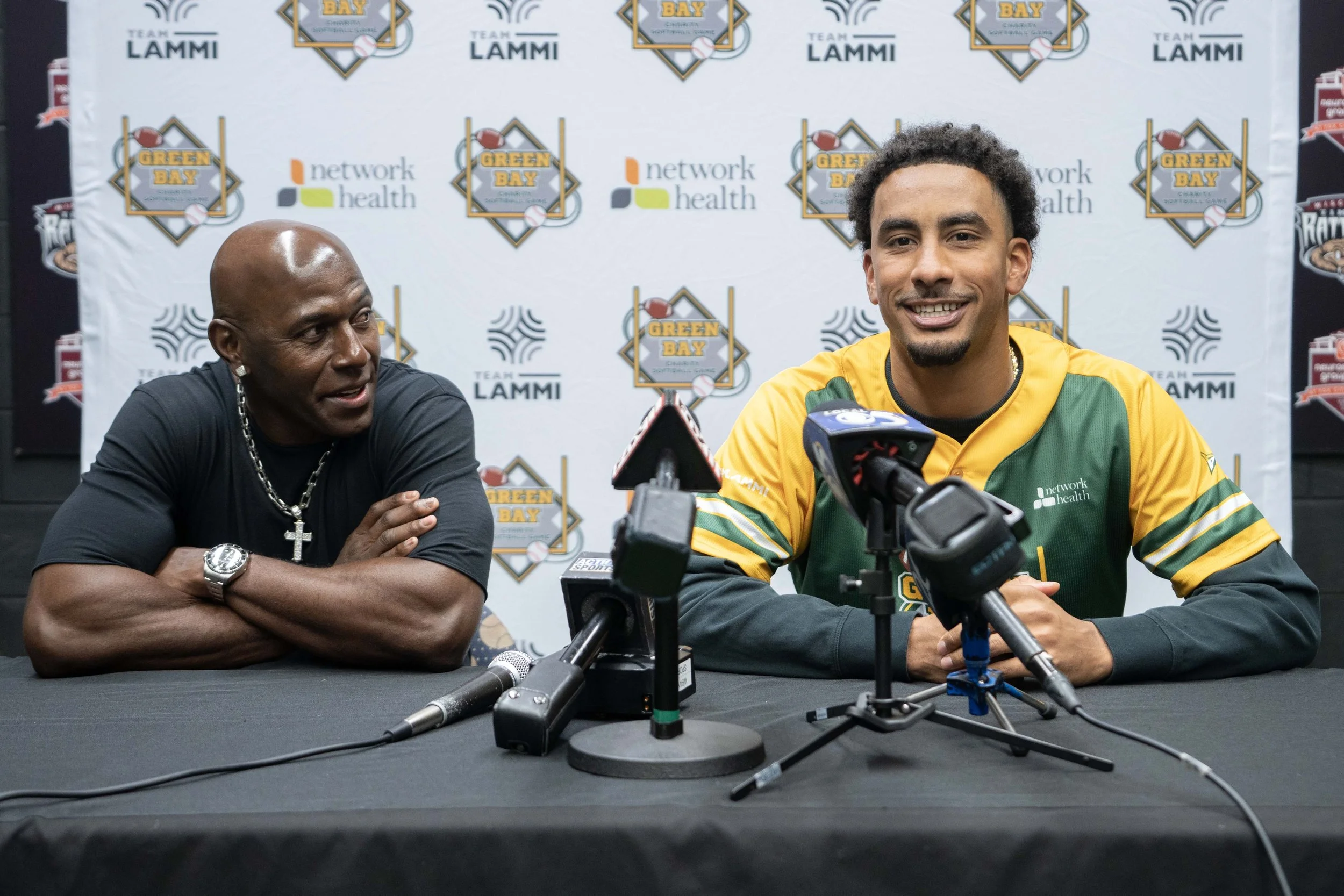 Two men at a press conference, one wearing a dark shirt with a chain and watch, the other in a green and yellow sports jacket, sitting at a table with microphones, with a backdrop featuring logos for Green Bay and Team Lammi.