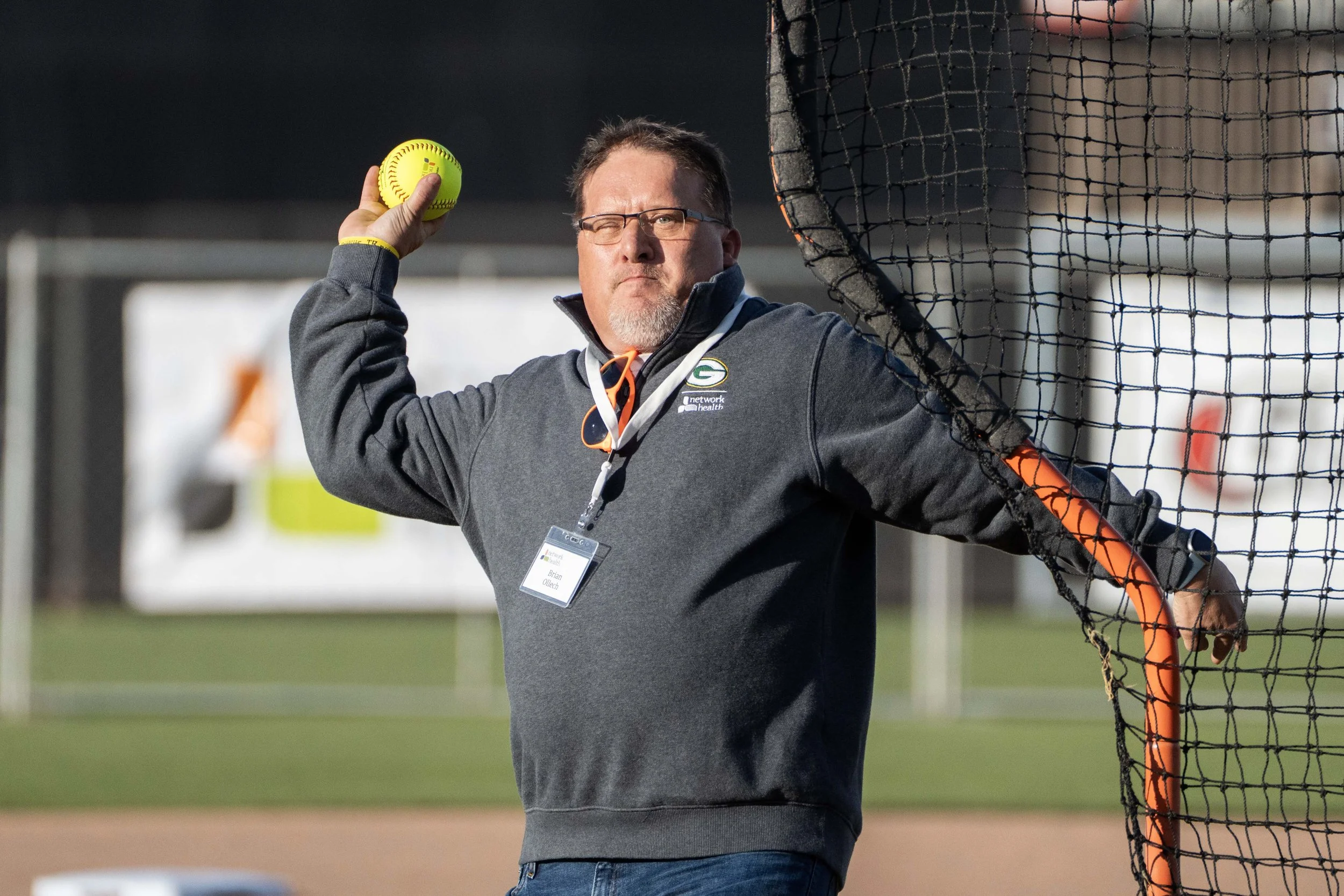 A man in a gray sweatshirt and glasses is standing on a baseball field, holding a yellow softball with his right hand, and resting his left arm on a black net fence.