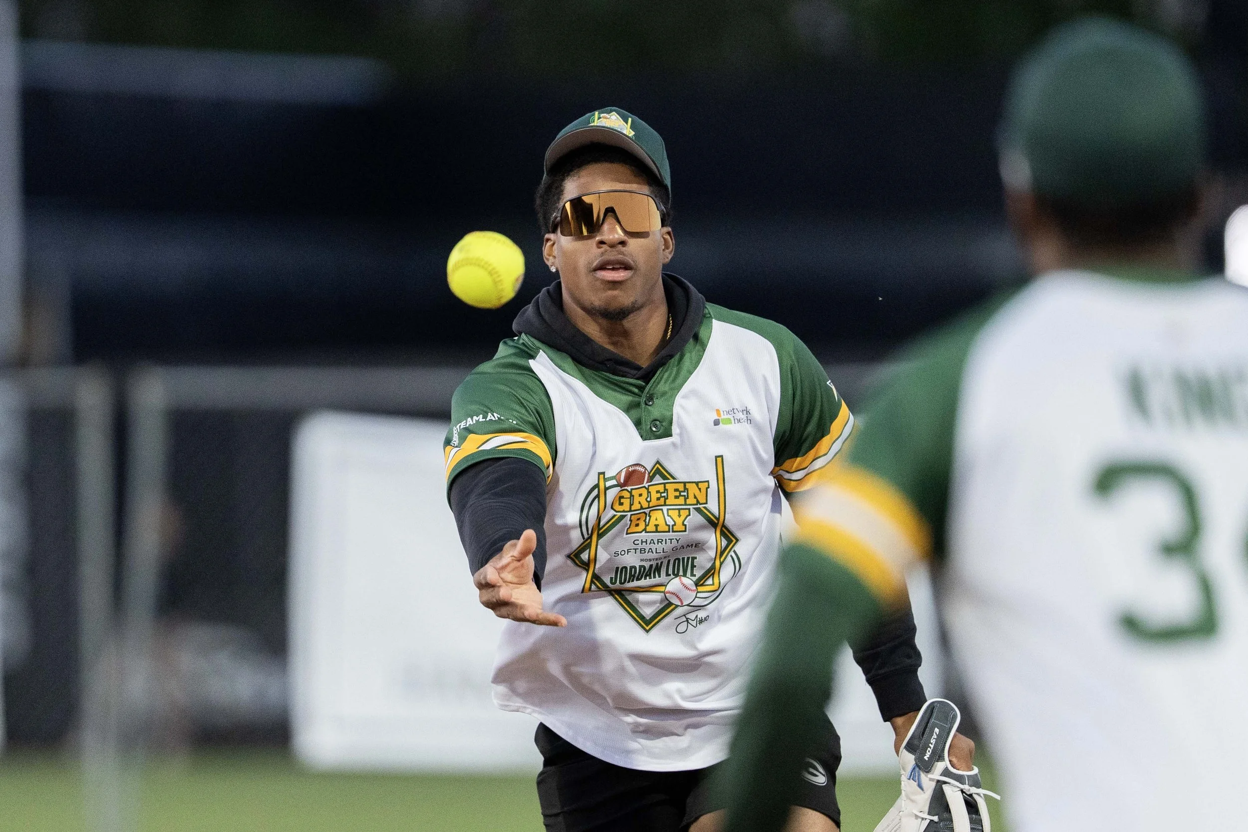 A young man in a Green Bay charity softball game shirt wearing sunglasses, a baseball cap, and a black hoodie. He is playing softball and is in motion, preparing to catch or throw the yellow softball ball.