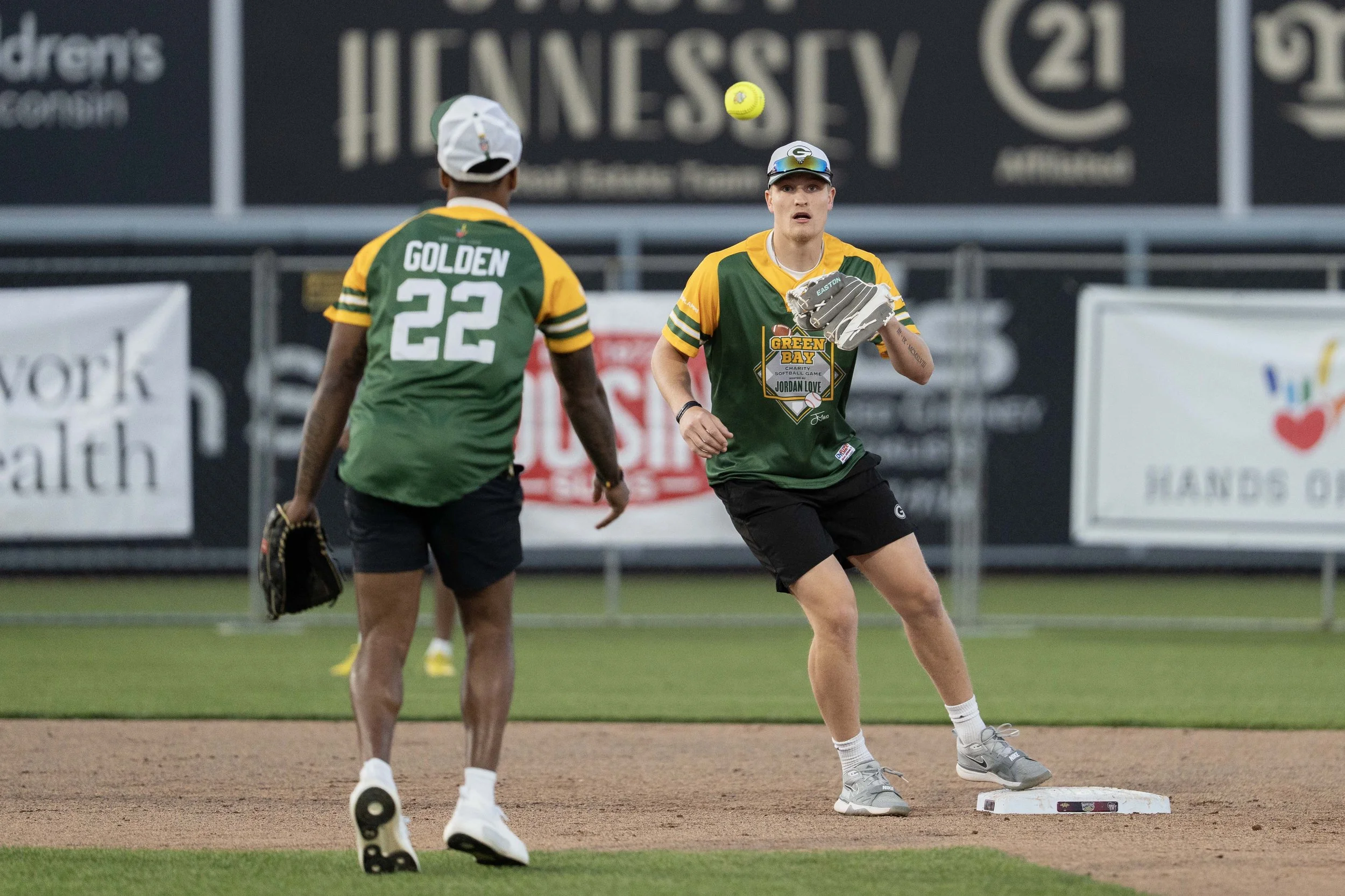 Two baseball players in green and yellow uniforms on a field, one wearing jersey number 22 and the other holding a glove, preparing to catch a ball.