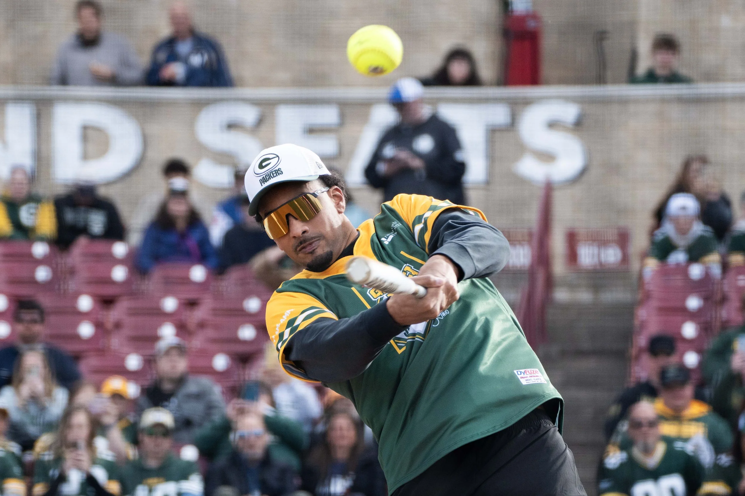 A baseball player in Green Bay Packers jersey and sunglasses hits a baseball with a bat during a game or practice, with a crowd watching in the background.