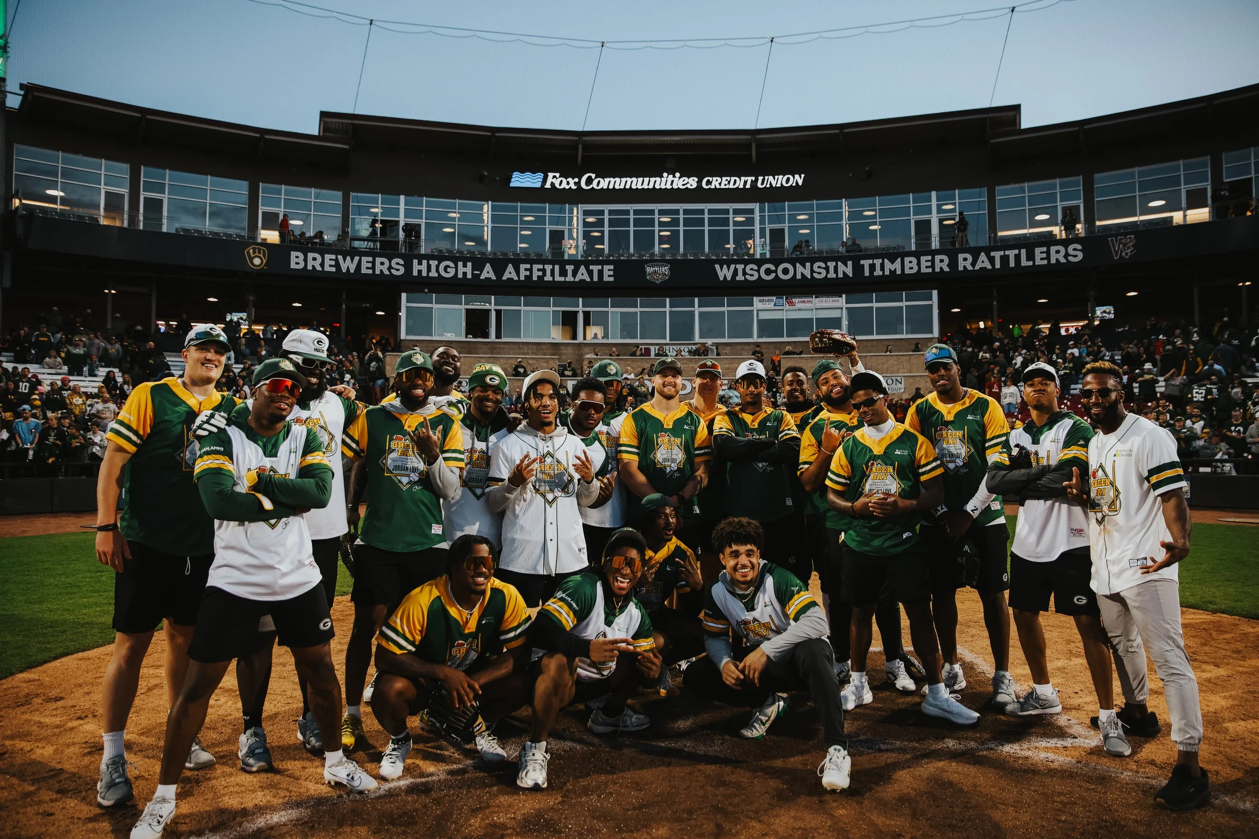 A group of baseball players in colorful uniforms and sunglasses celebrating on a baseball field after a game, with a stadium and spectators behind them.