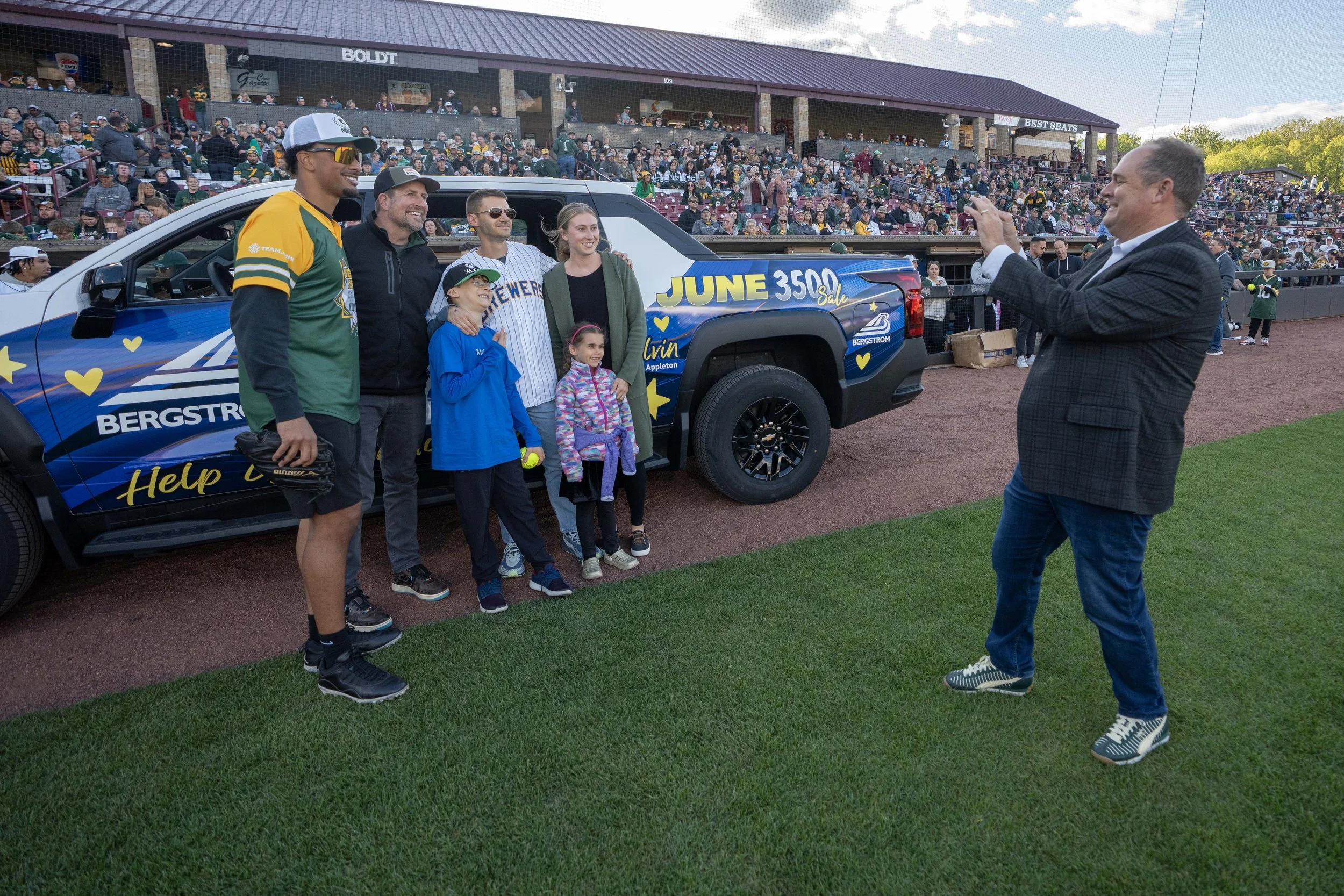 A group of people, including children and adults, is taking a photo with an adult man on a baseball field, with a large crowd of spectators in the bleachers behind them. The group is standing next to a blue pickup truck decorated with advertisements,