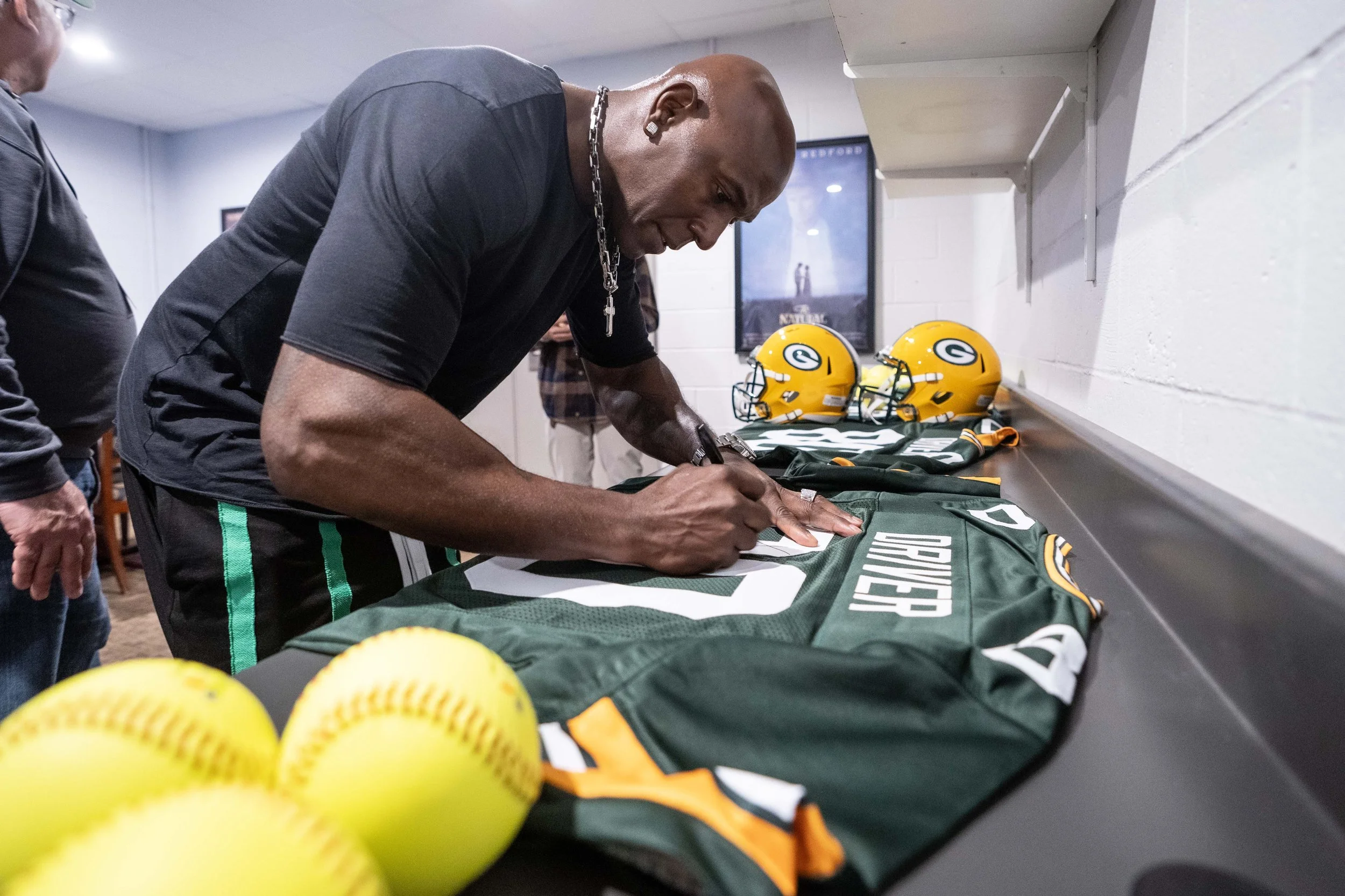 A man signing Green Bay Packers jersey in a locker room with football helmets and footballs nearby.