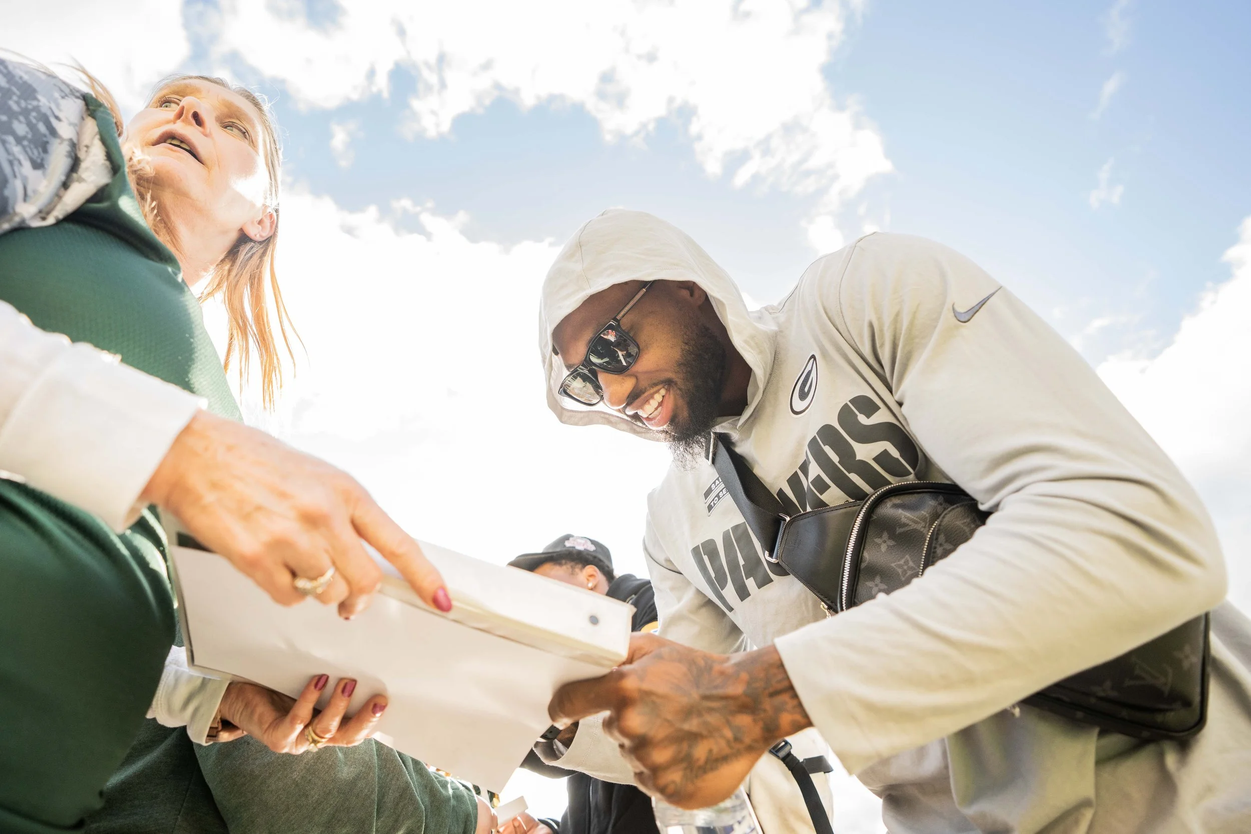 A man wearing sunglasses and a hoodie, smiling, signs an autograph for a woman with long hair, wearing a green jacket, against a bright sky with clouds.