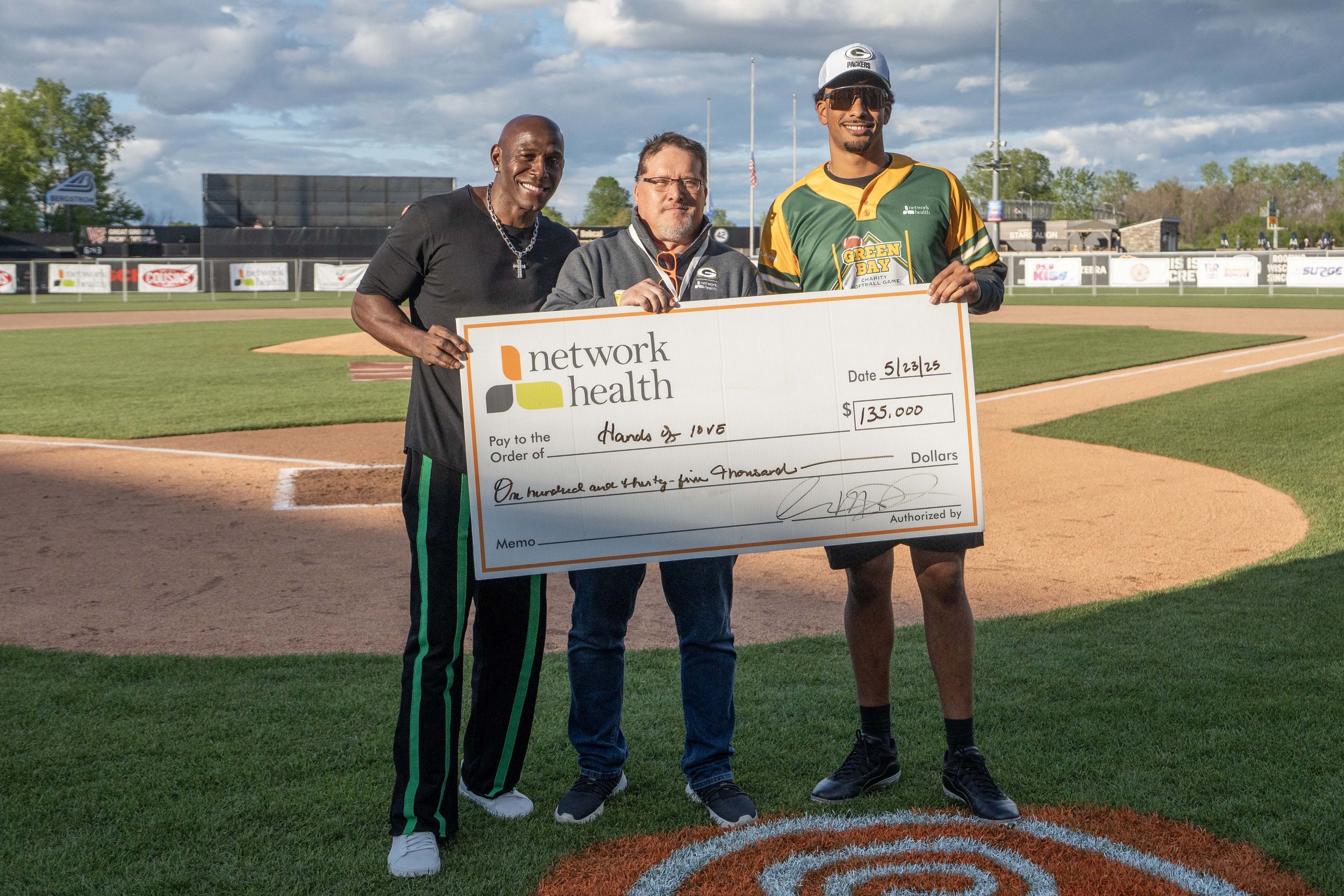Three men standing on a baseball field holding a large check for $135,000 made out to 'Hands & 105.' The man on the right is wearing a green and yellow football jersey, shorts, sneakers, sunglasses, and a cap. The man in the middle is wearing glasses