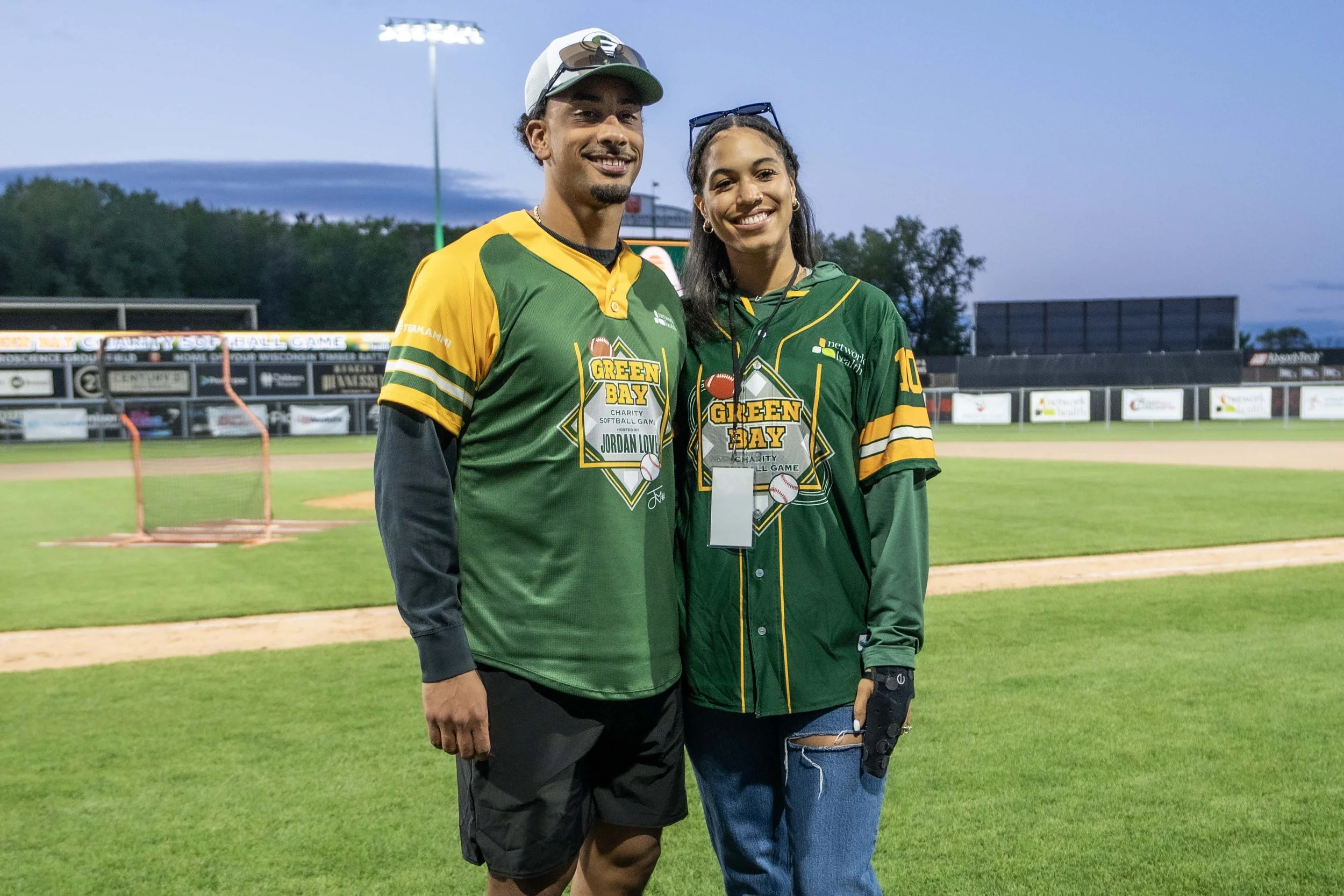 Two people standing on a baseball field at dusk, wearing green and yellow charity softball tournament jerseys with 'Green Bay' logos. They are smiling, with one wearing a cap and sunglasses and the other with glasses on her head and a glove on her ha
