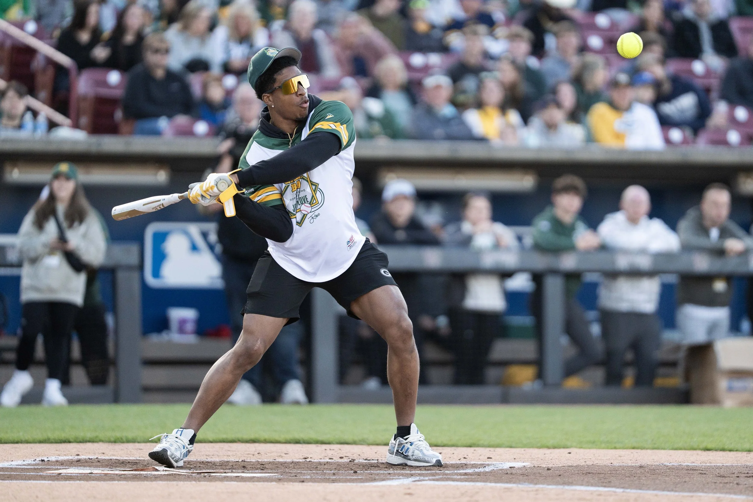 A baseball player in a green and white uniform hitting a yellow softball during a game, with spectators watching.