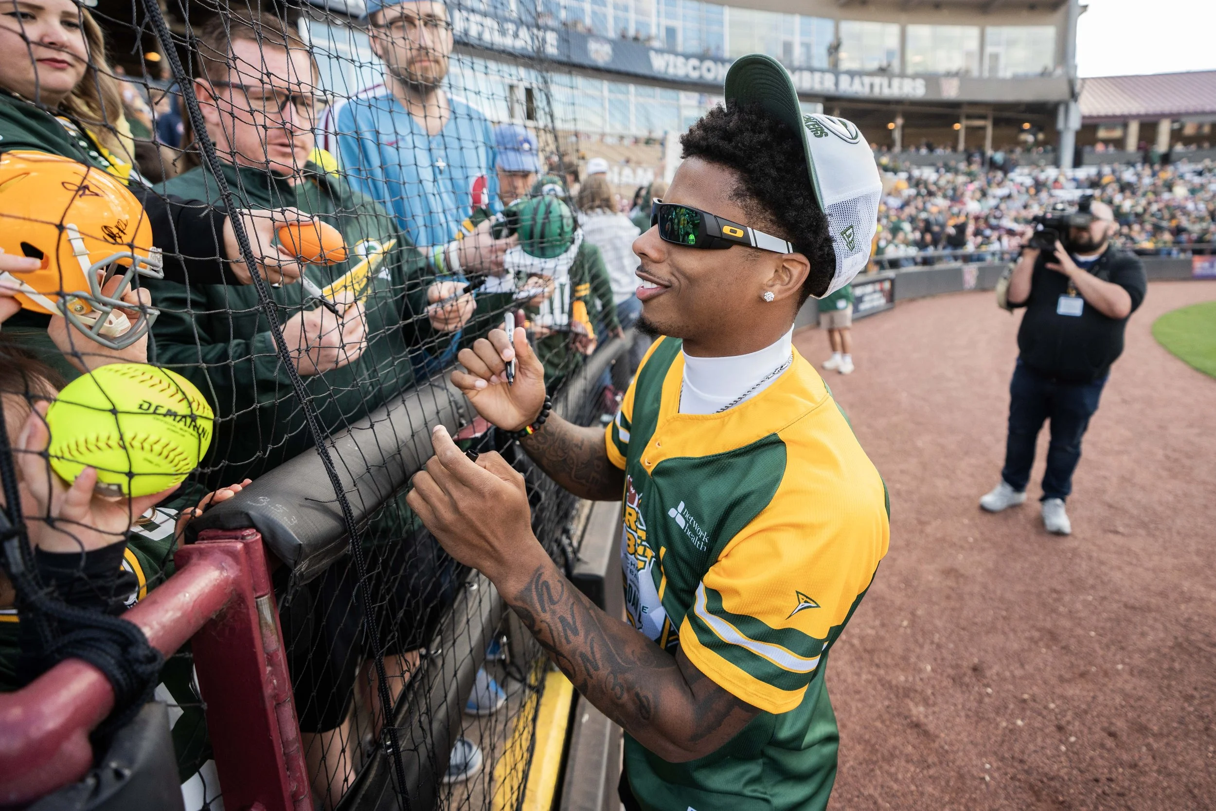 A professional baseball player signing autographs for fans behind a protective fence at a stadium. The player is wearing sunglasses, a cap, and a green and yellow uniform, with tattoos visible on his arms. Fans are holding out baseballs and phones fo