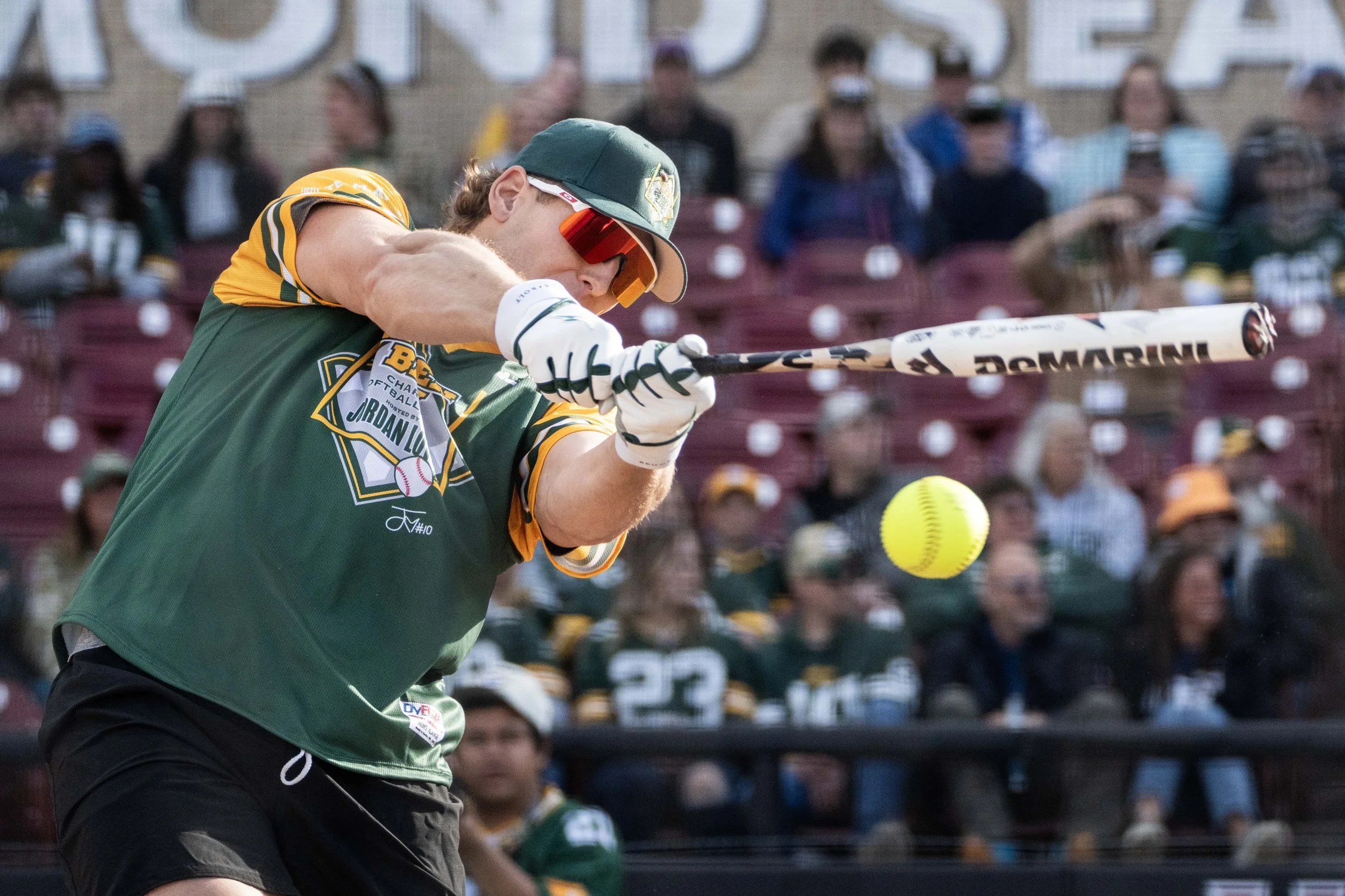 A baseball player in a green and yellow uniform hits a softball with a bat during a game, with spectators watching in the stands in the background.