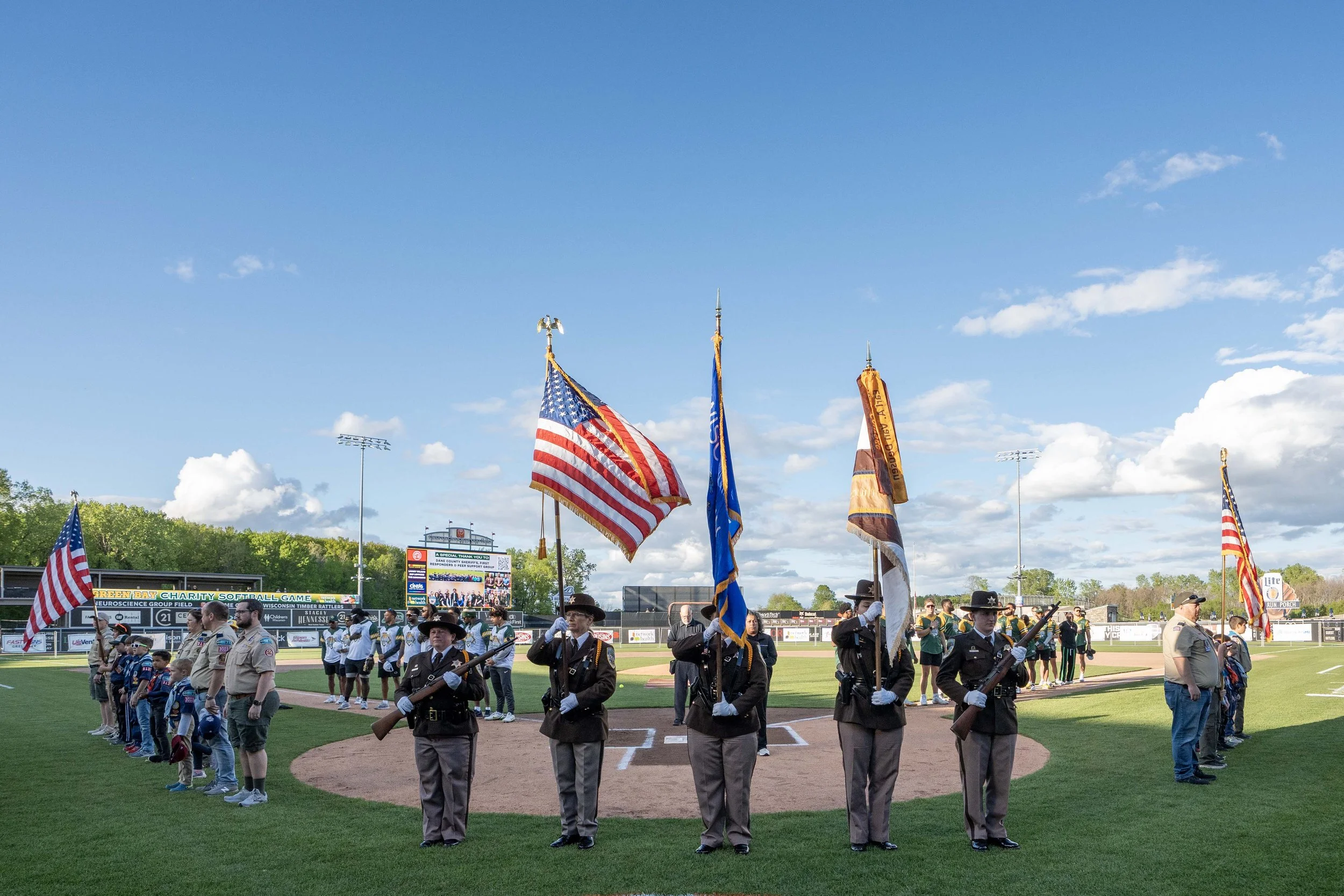 People standing on a baseball field for a pre-game ceremony, holding flags including American flags and other banners, with stadium seating and scoreboard in the background on a clear day.