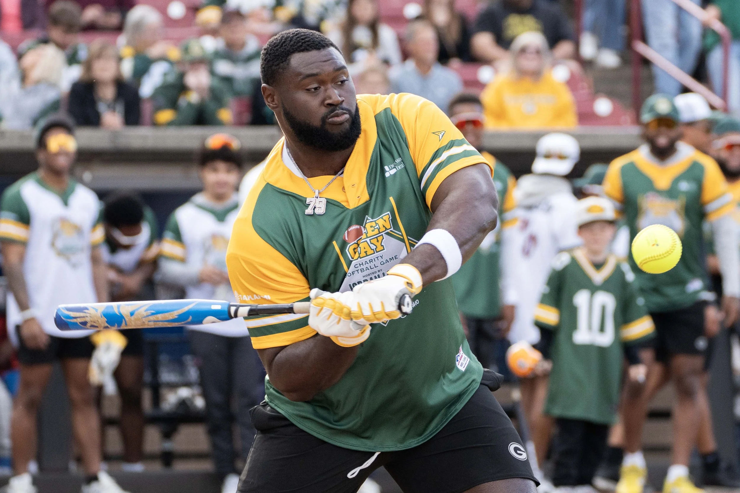 Man in green and yellow football jersey hitting a softball with a bat during a game, with fans watching in the background.
