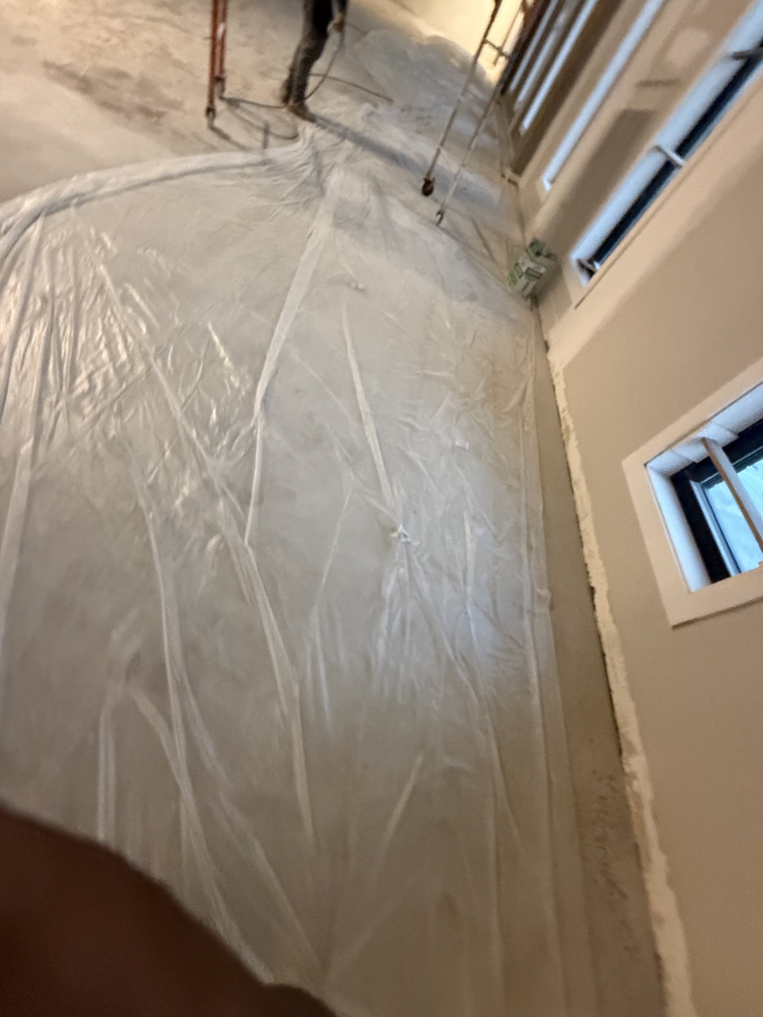 Room under construction with beige walls, two small windows, and a protective plastic sheet on the floor. Construction tools and materials are visible.