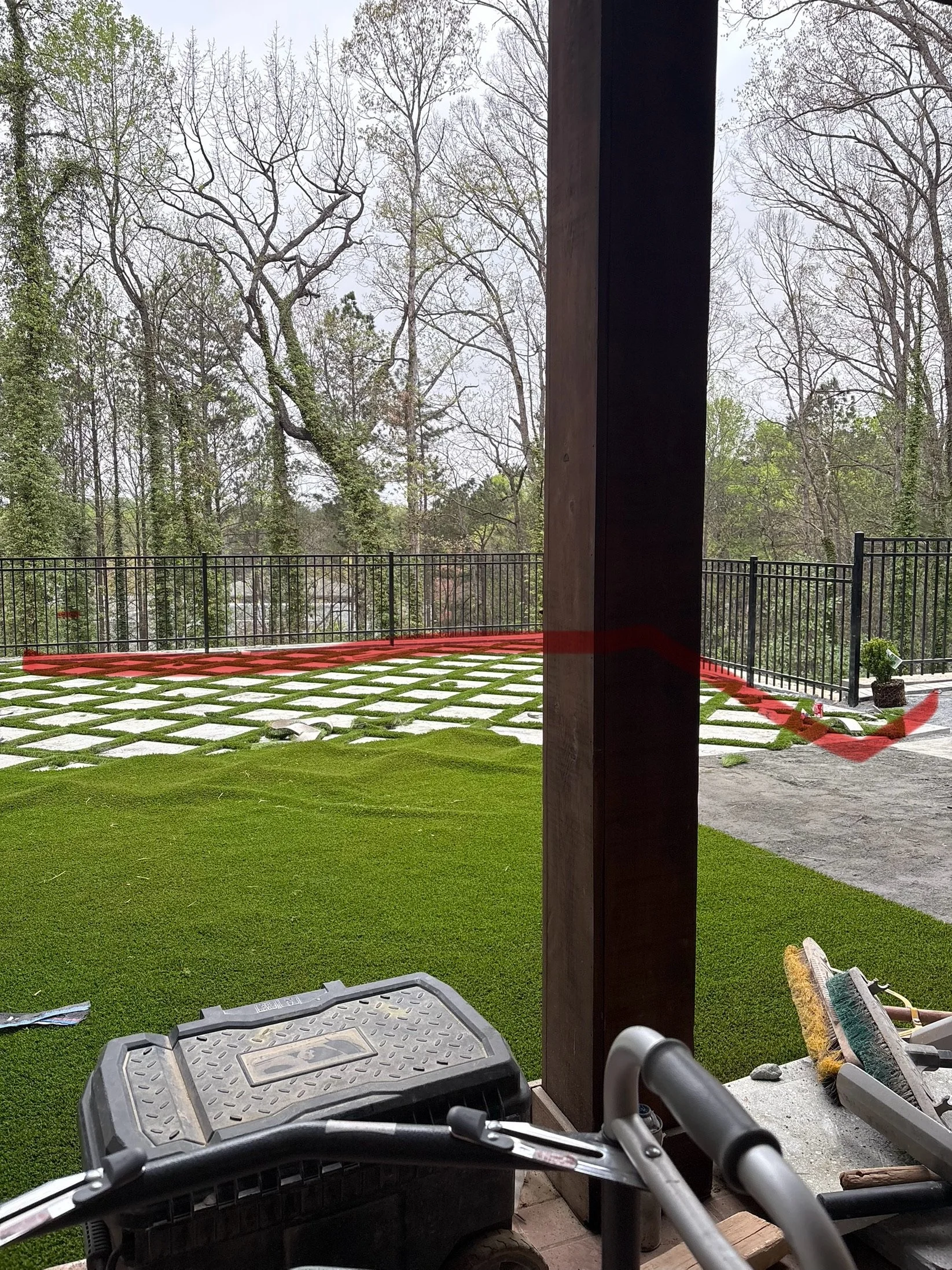 View of a backyard with a partially completed grass and stone patio, black metal fence, and bare trees in the background, seen from inside a covered porch with construction tools in the foreground.