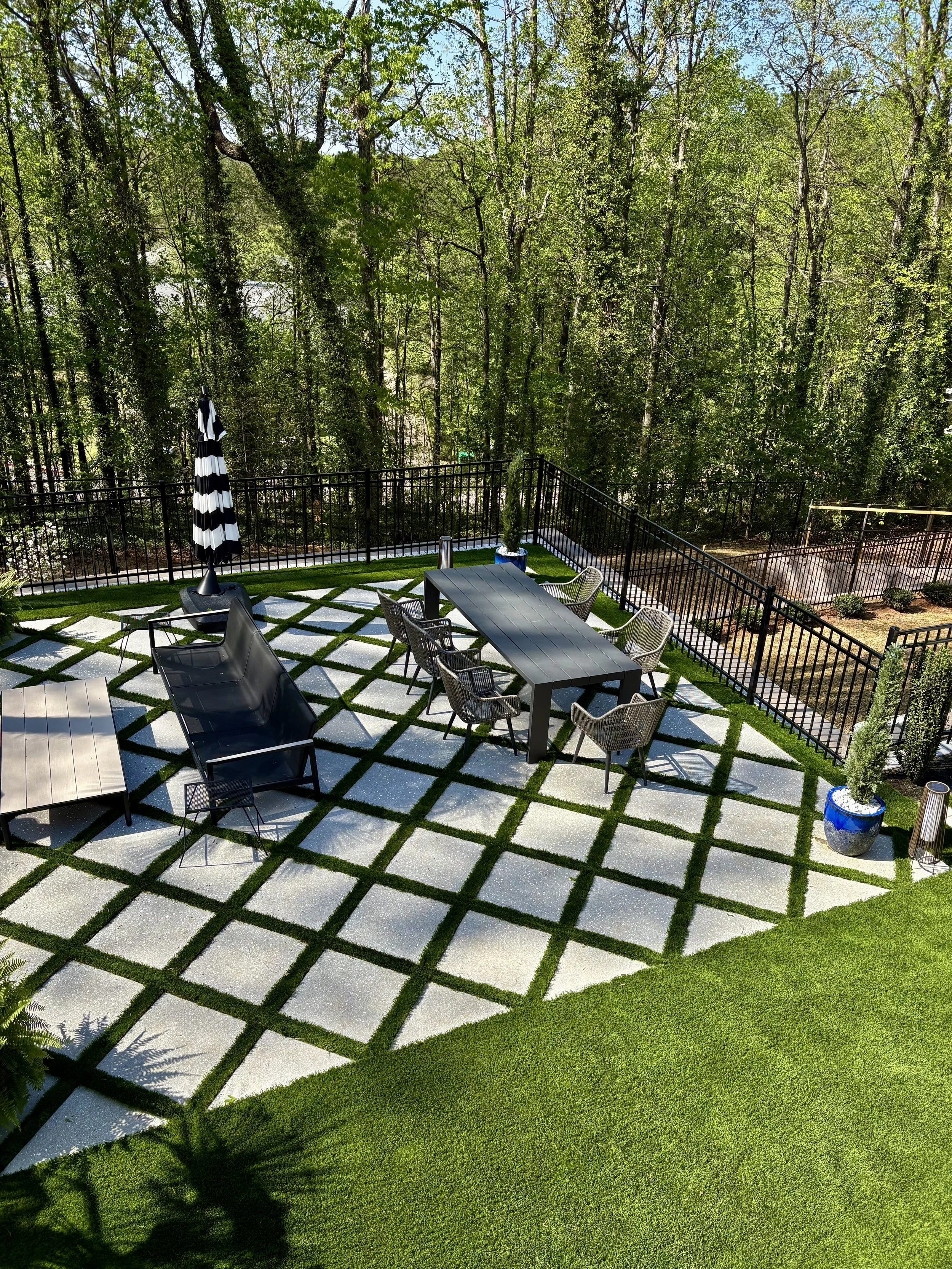 An outdoor patio area with a modern black dining table and six chairs, a large black umbrella, a black lounge chair, and decorative potted plants, surrounded by a black metal fence and lush green trees.