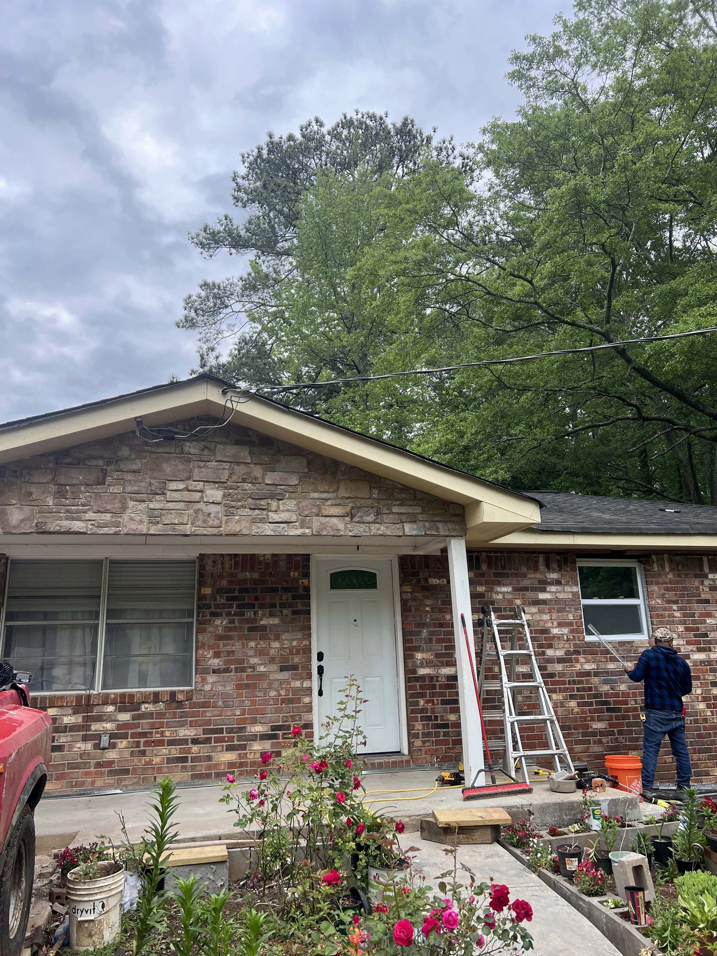 A person is power washing the exterior wall of a brick house in a garden with pink flowers, a ladder, a lawn chair, and gardening tools nearby.