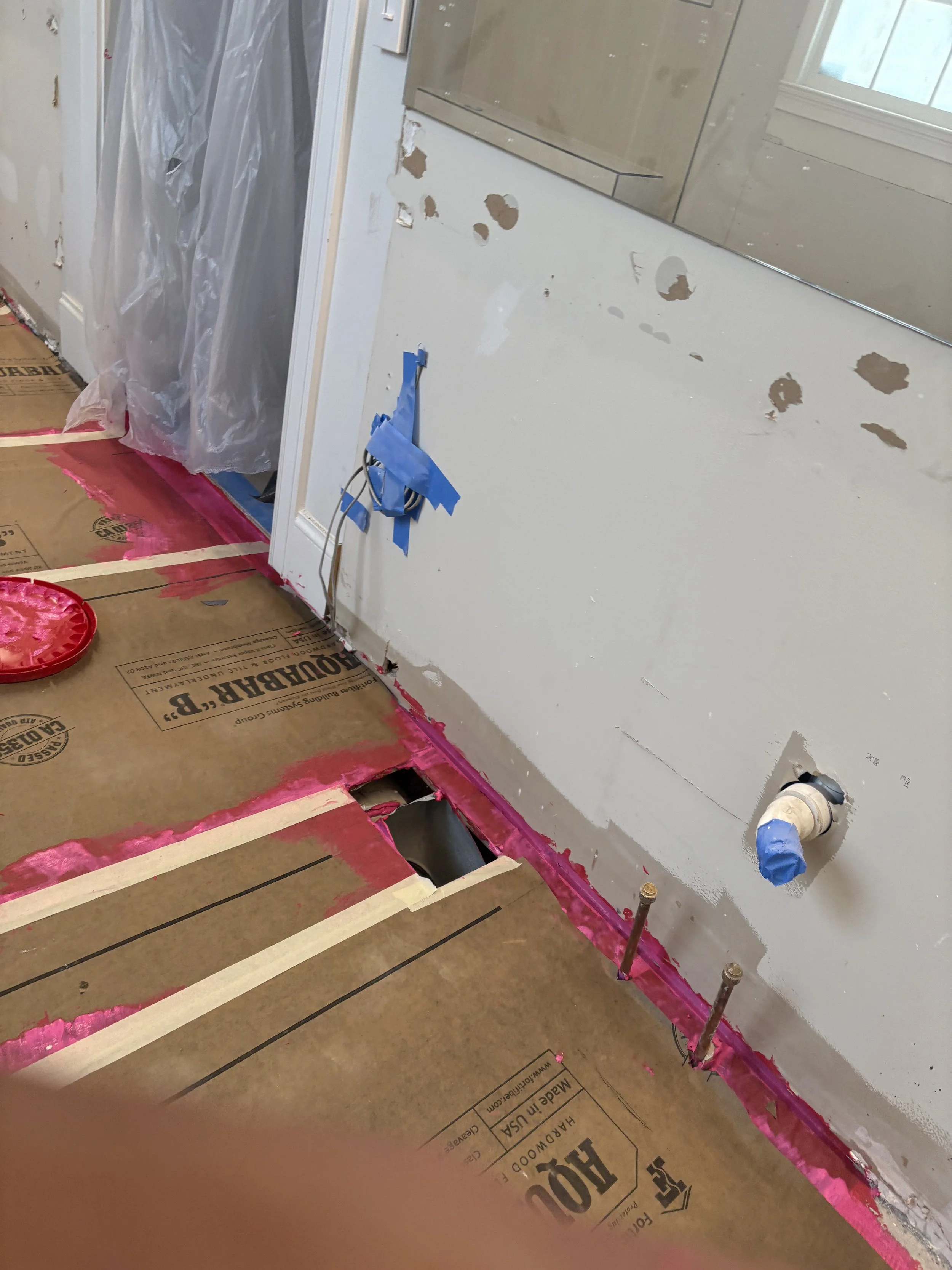 A room under renovation with drywall and plumbing work in progress. The floor is covered with cardboard for protection, and there are pink sealant lines along the edges. A window is visible, and there are some holes in the wall for electrical wiring 