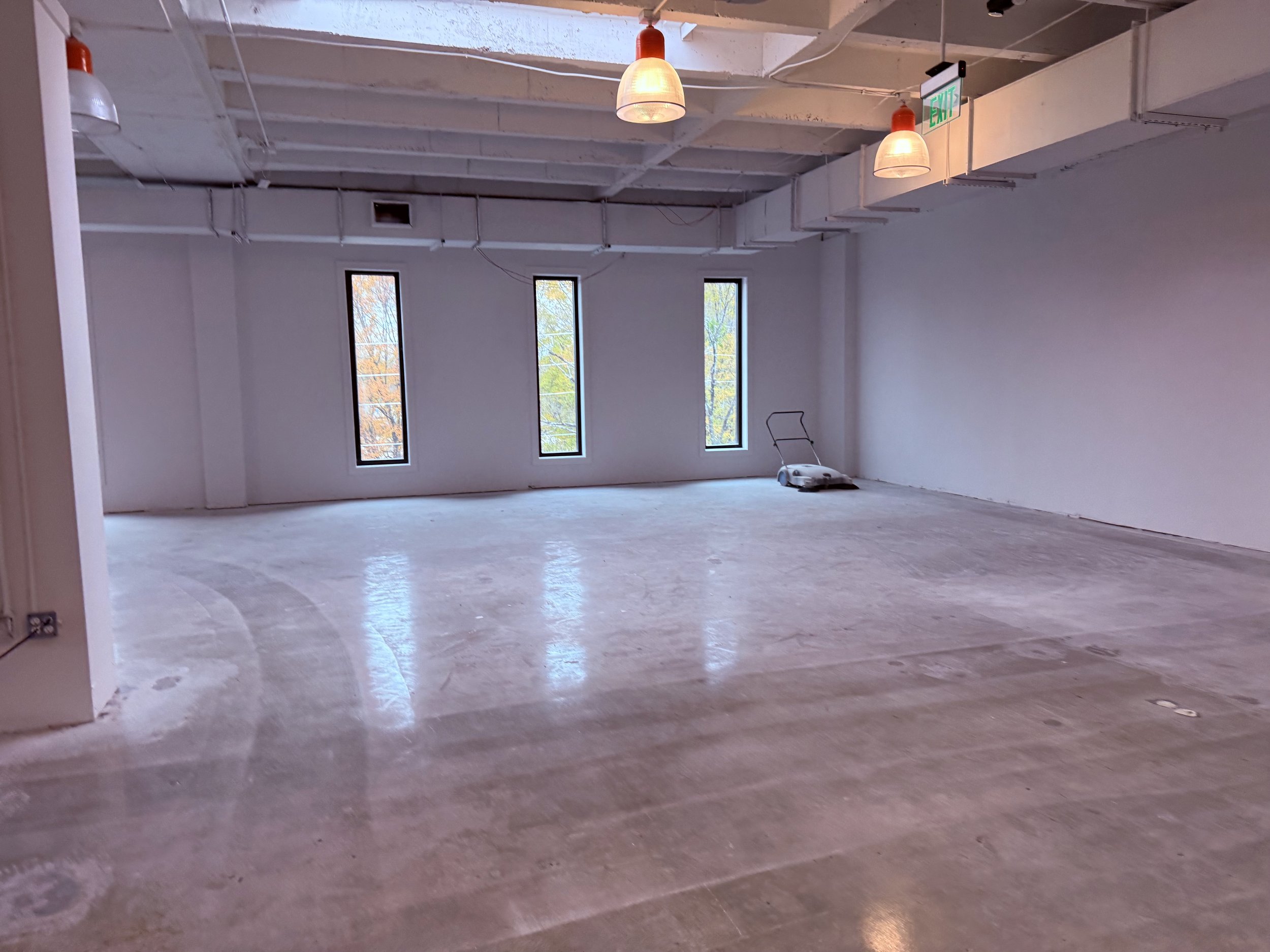 Empty room with three tall, narrow windows, hanging lights, and a floor polishing machine in the corner.