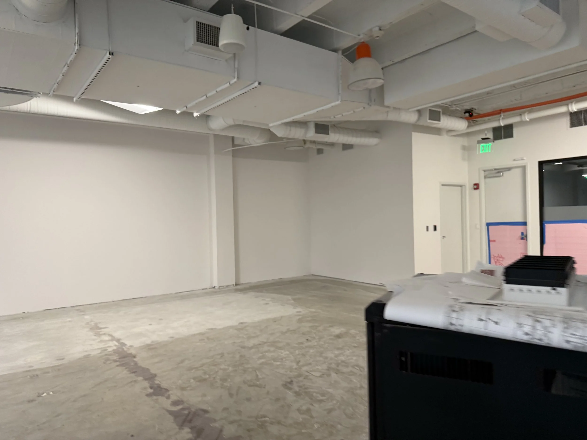 Empty commercial space with exposed ceiling pipes, a concrete floor, and white walls. An exit door with an illuminated green exit sign is visible on the right, and there are some construction materials on a table in the foreground.
