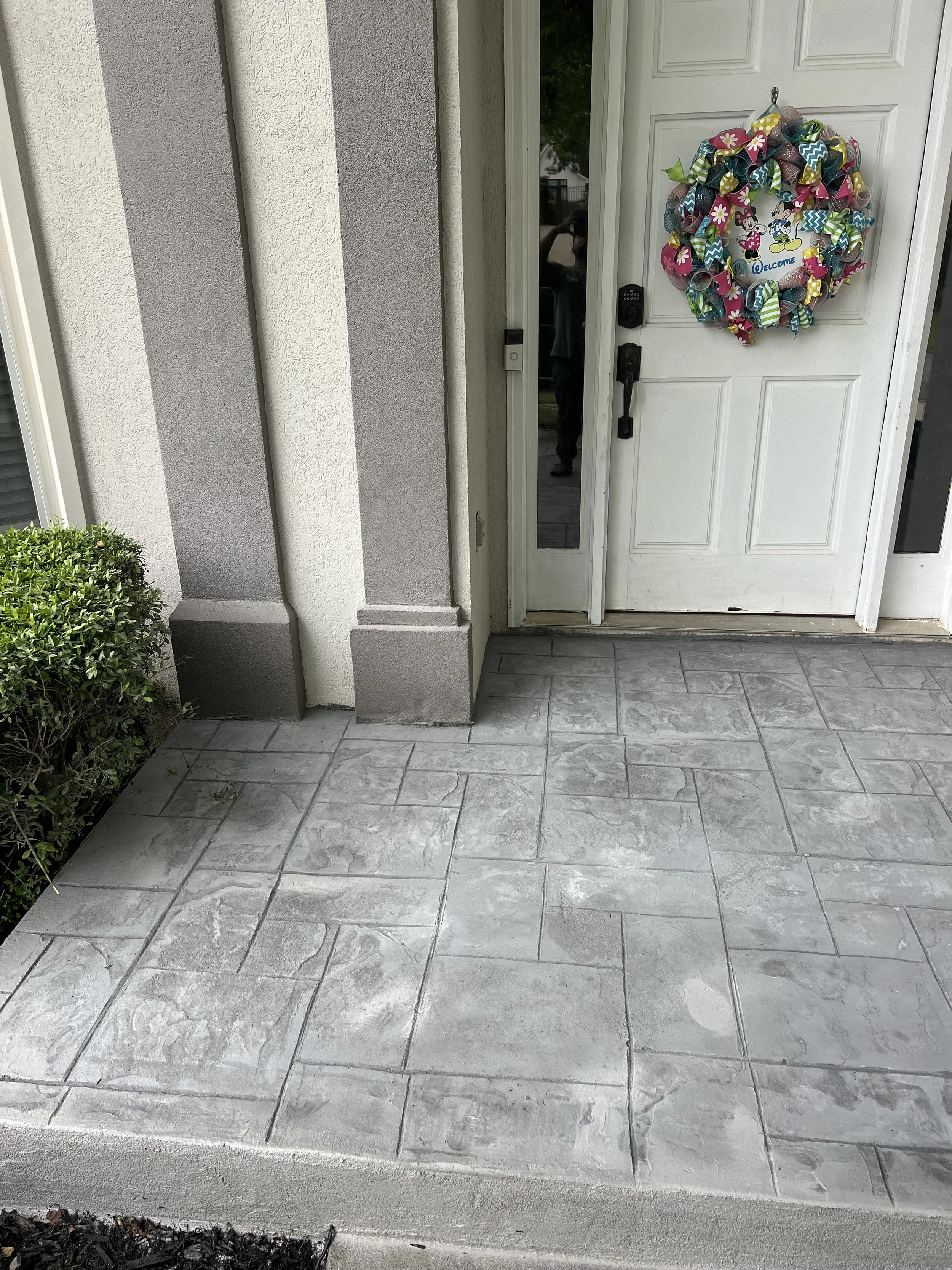 Front porch area of a house with a white door decorated with a colorful Minnie Mouse themed wreath and a small green shrub to the left.