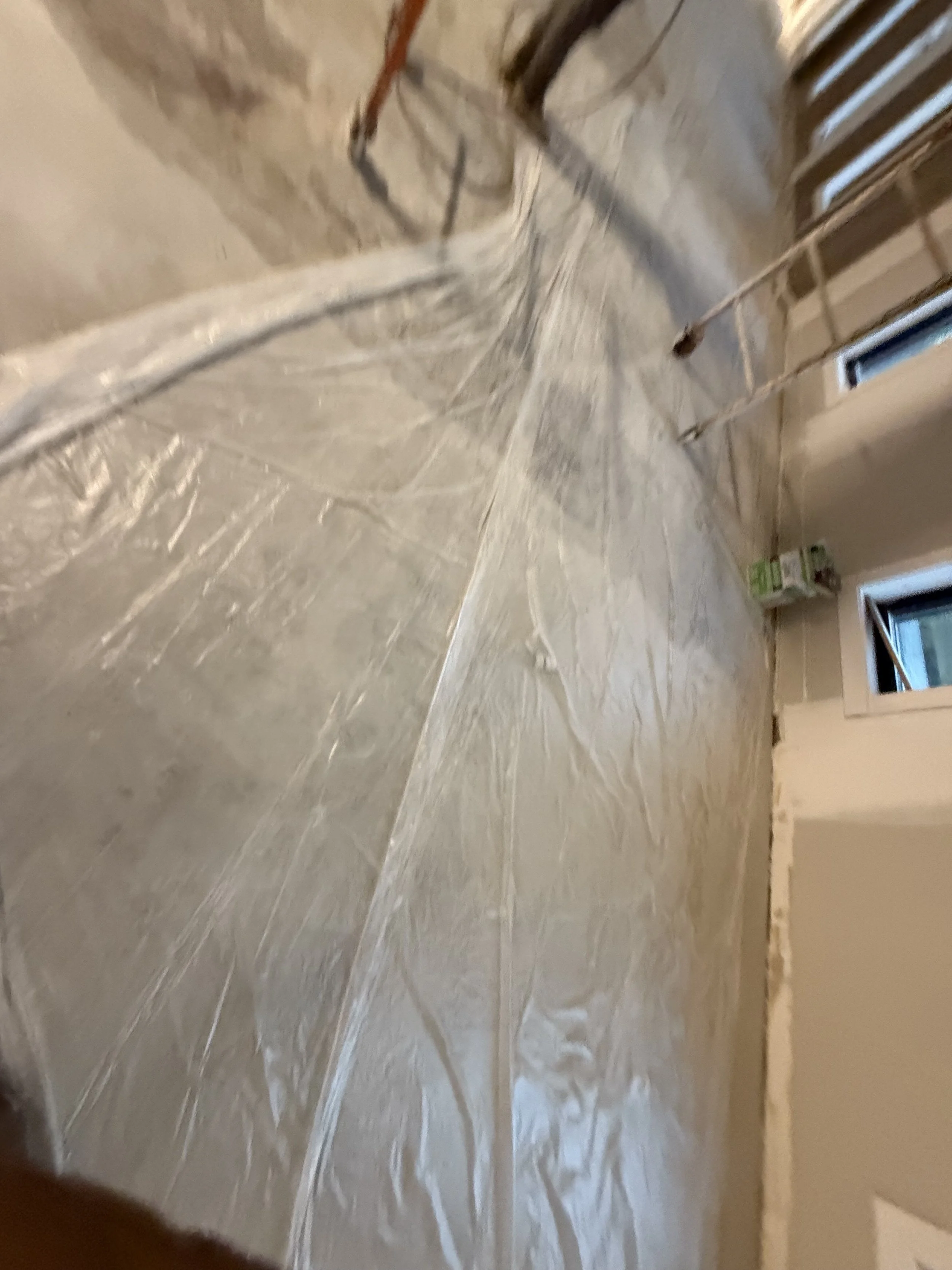 A room under renovation with plastic sheeting on the floor, around some pipes, and a partially unfinished wall with visible wiring and a window.