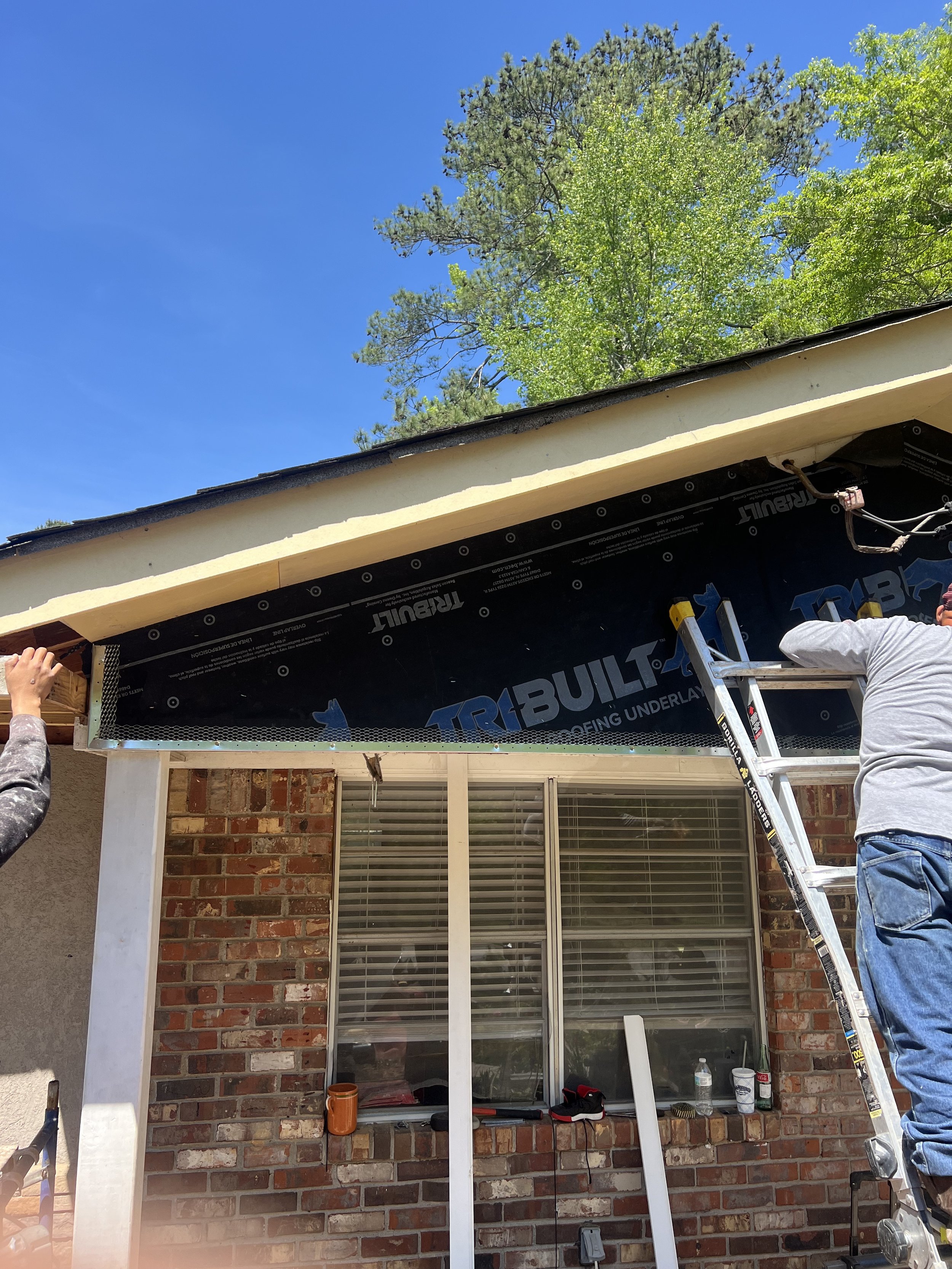 Construction workers installing or repairing the soffit and fascia on the exterior of a brick house, with a ladder leaning against the wall, while a bright blue sky and trees are visible above.