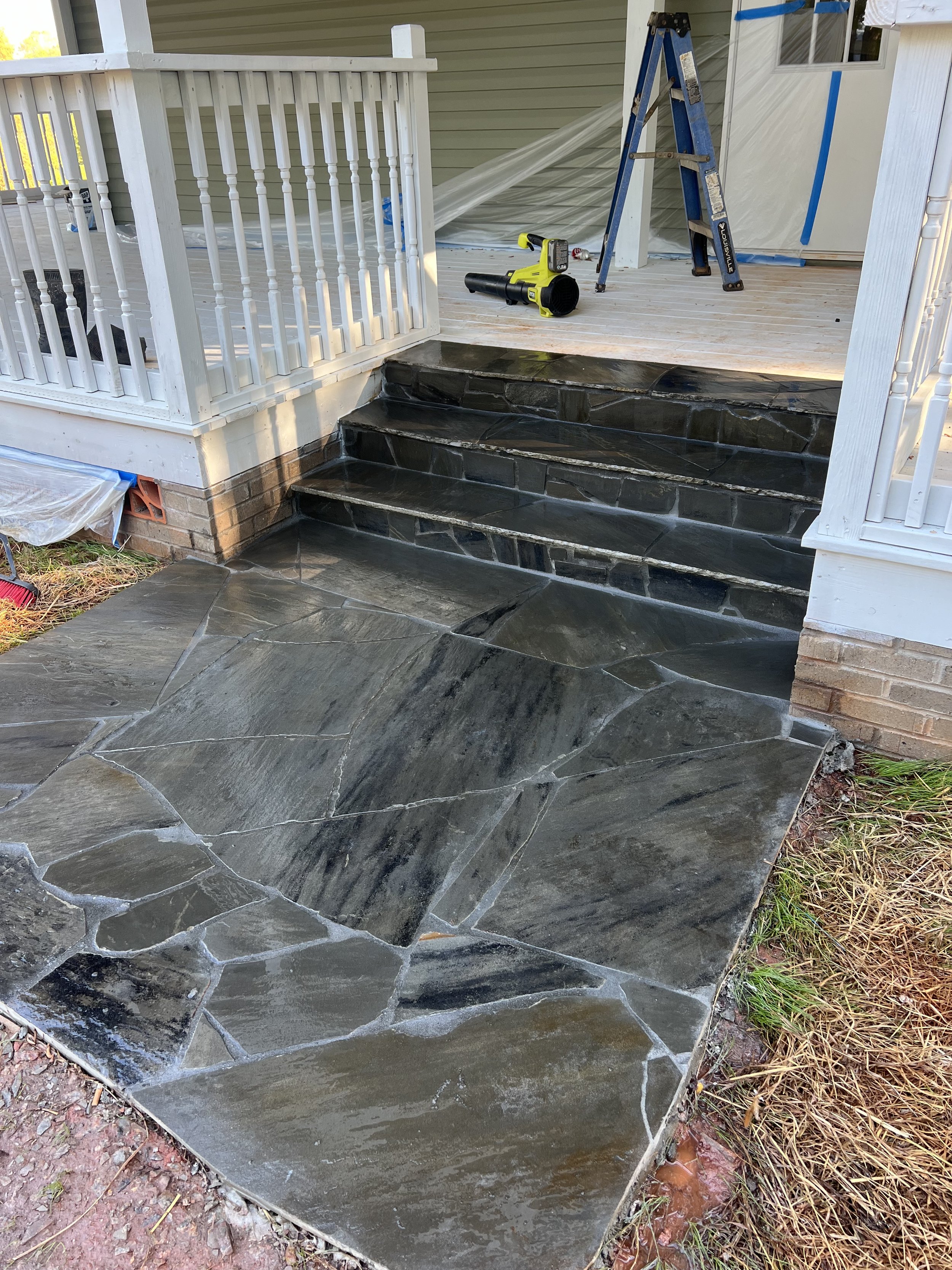 Stone stairs leading to a porch with a white railing, tools like a ladder, a leaf blower, and a drill are placed on the porch, and the stairs are being cleaned or sealed.