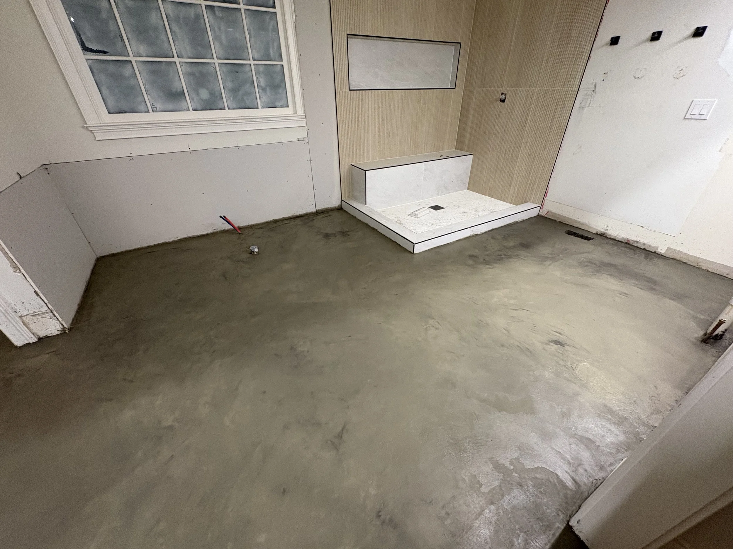 Room under renovation with unfinished concrete floor, a window, and partially installed tiles around what appears to be a shower area.