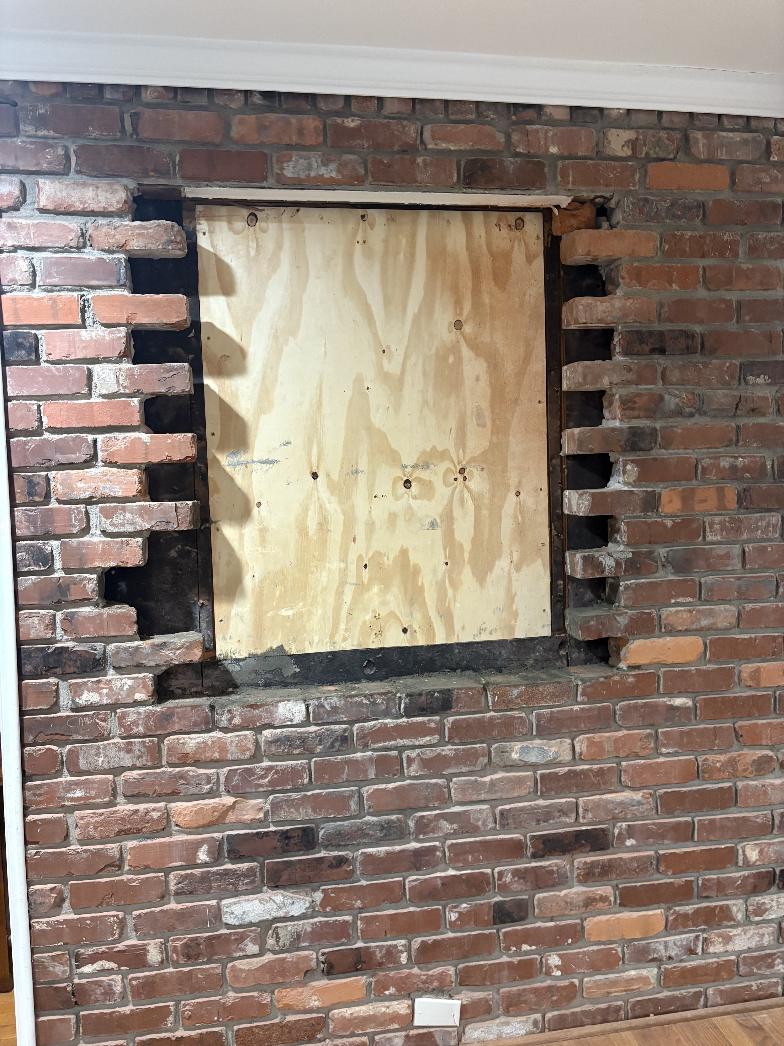 Brick wall with a missing section filled with plywood board covering an opening.