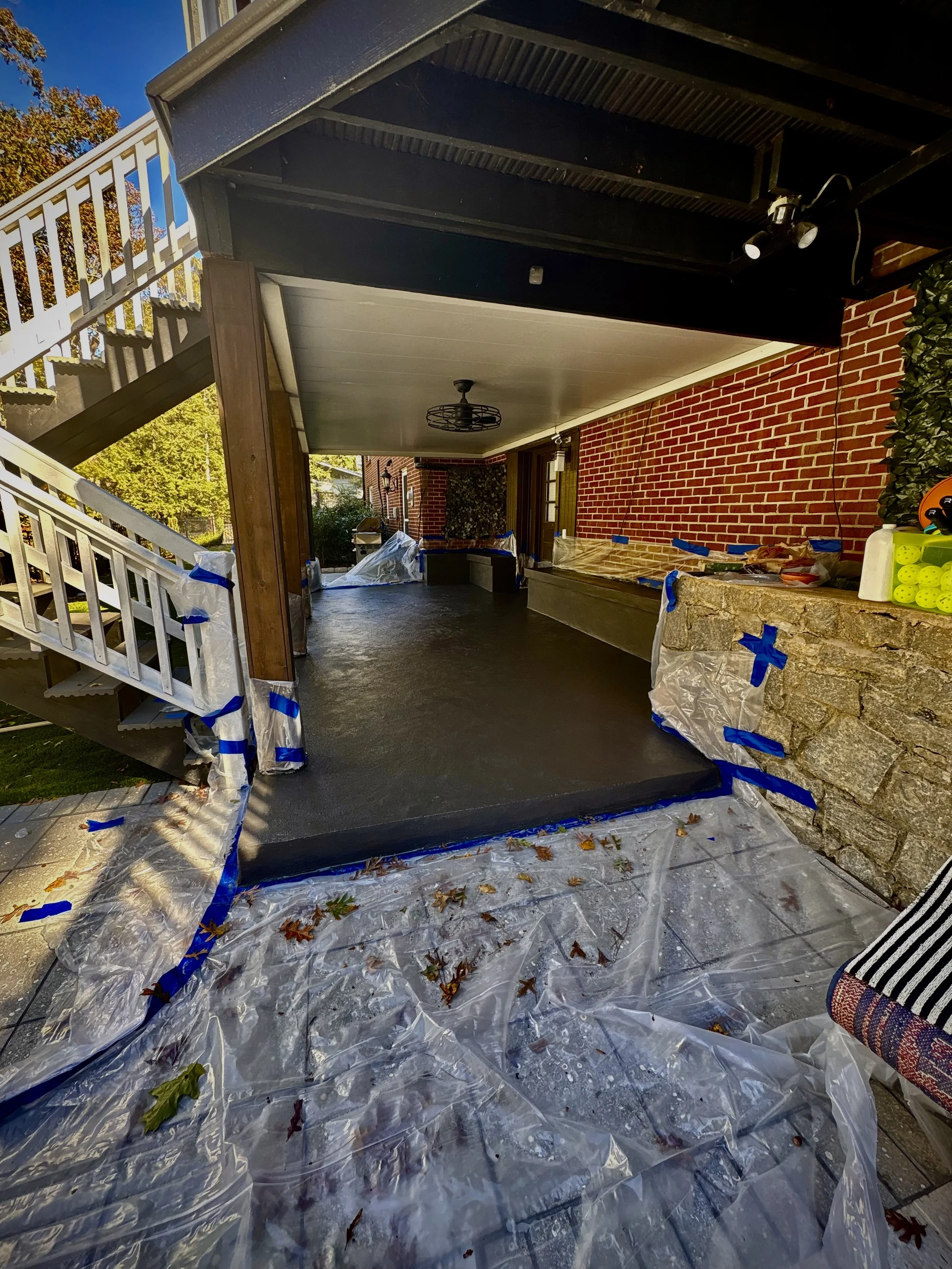 A covered porch area with a freshly poured concrete floor, surrounded by plastic sheeting and blue painter's tape for protection during work, with a brick wall and outdoor staircase in the background.
