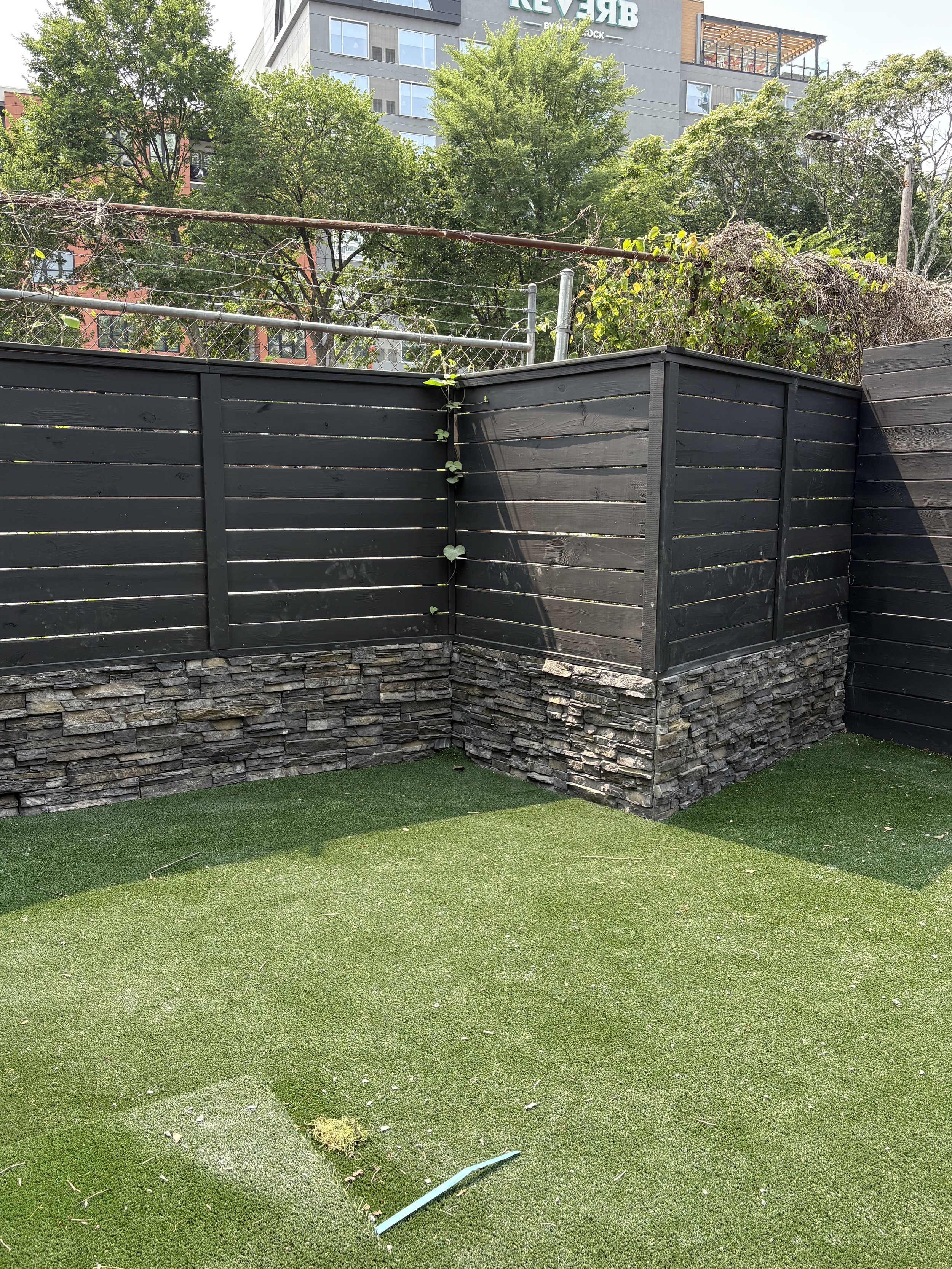 Empty outdoor area with artificial grass, a black wooden fence with a stone base, and trees in the background. A building with the word 'REVER' partially visible is in the distance.
