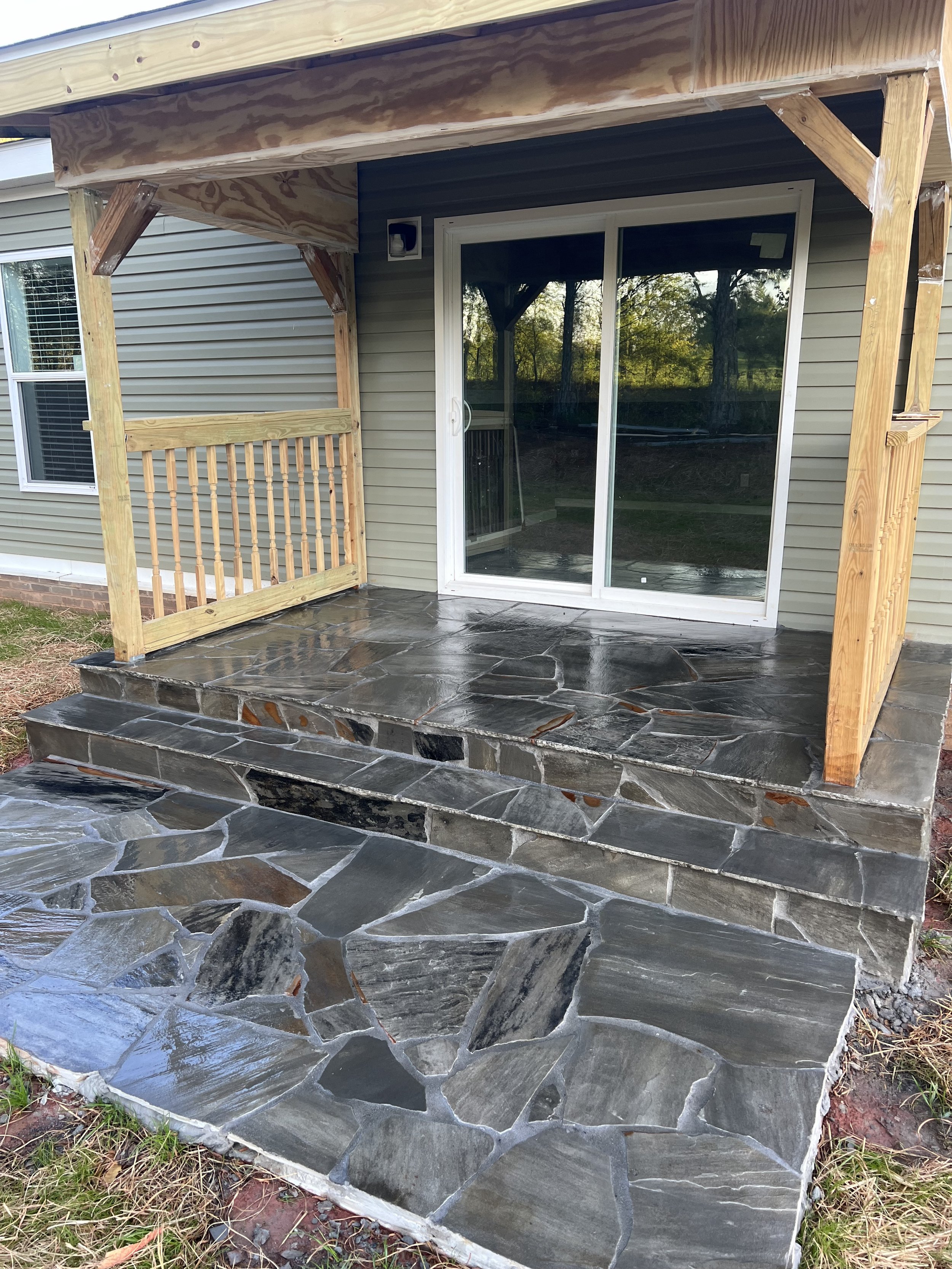 Stone patio with a couple of steps leading up to a sliding glass door of a house, with a small porch area and wooden railing in construction.