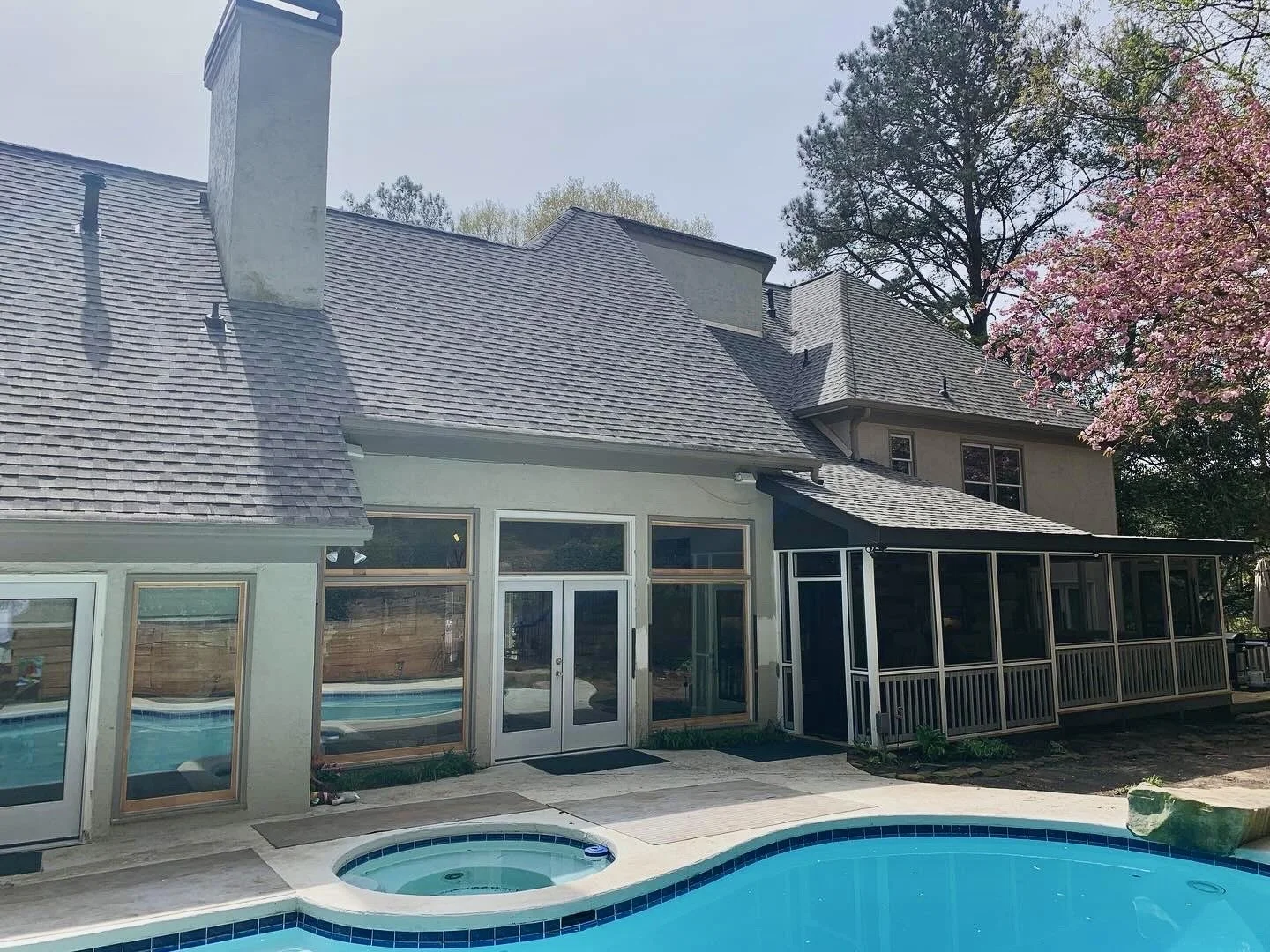 Backyard view of a house with a swimming pool, a hot tub, and a screened porch, surrounded by trees in springtime.