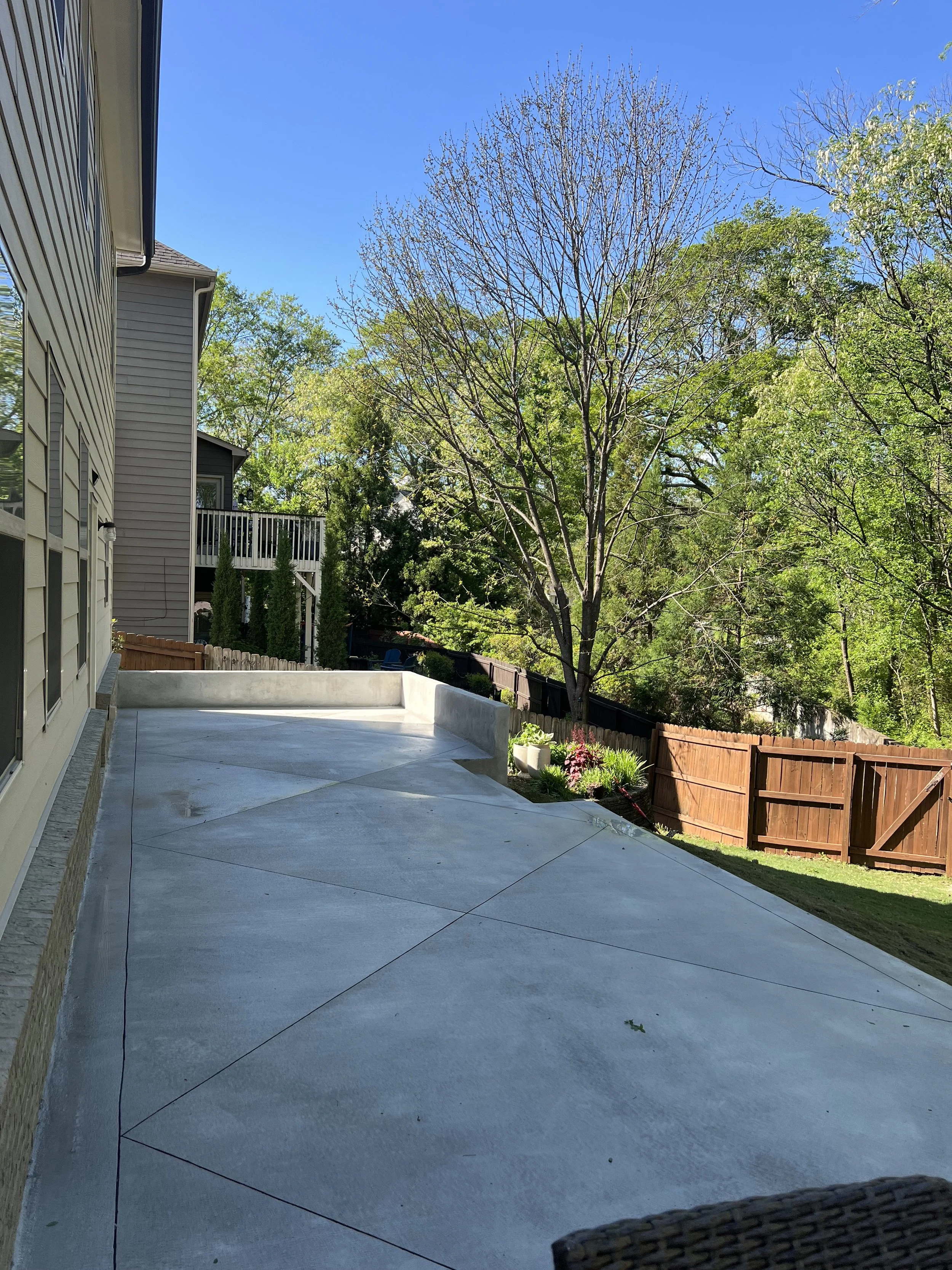 View of a backyard patio with a concrete surface, adjacent to a house with beige siding, a railing, a garden with flowers, and a wooden fence surrounding the yard. There are trees with some leaves and some without, under a clear blue sky.