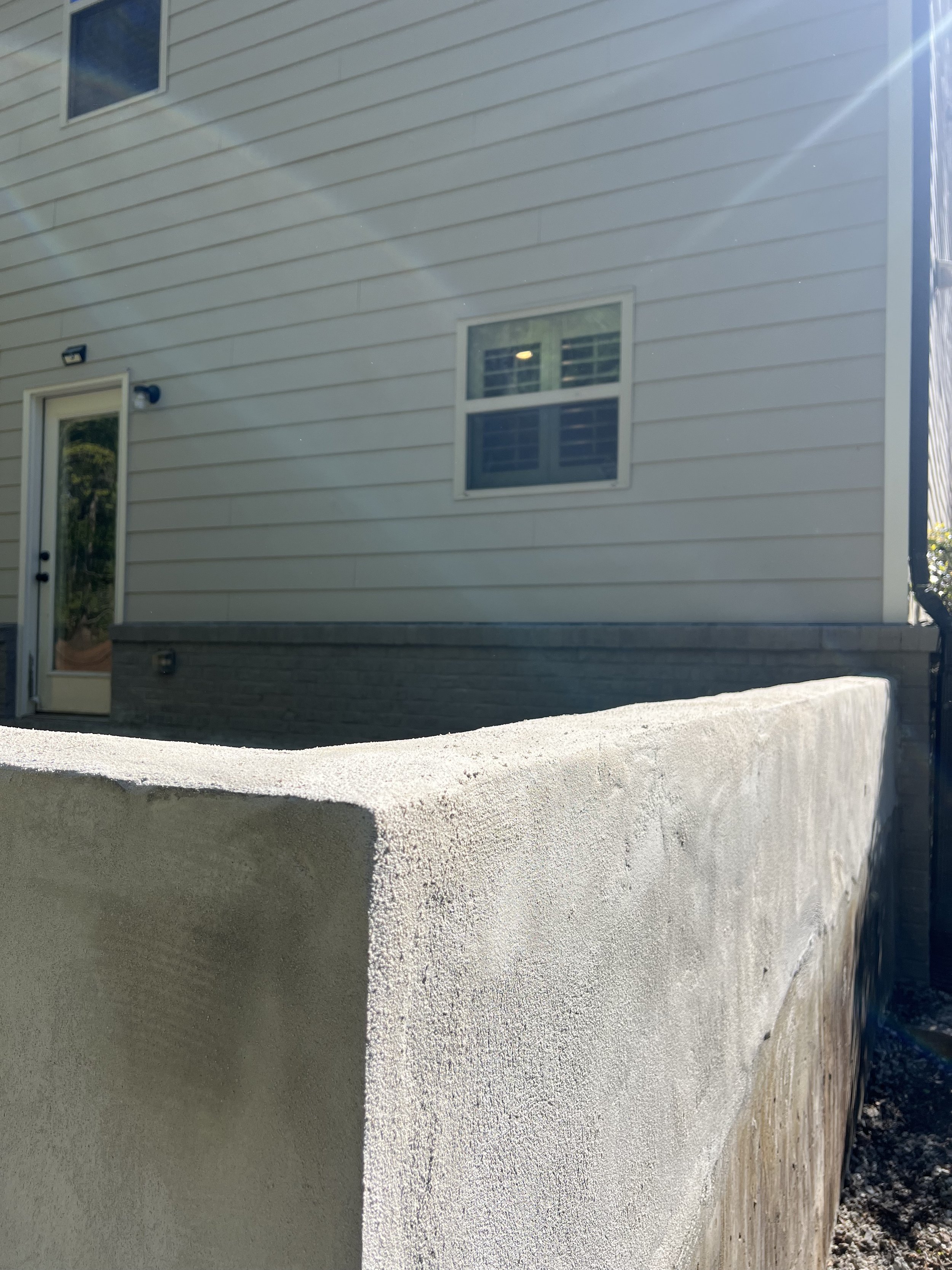 Exterior view of a house with beige horizontal siding, two windows, a door with glass panels, and a protective wall or barrier in the foreground.