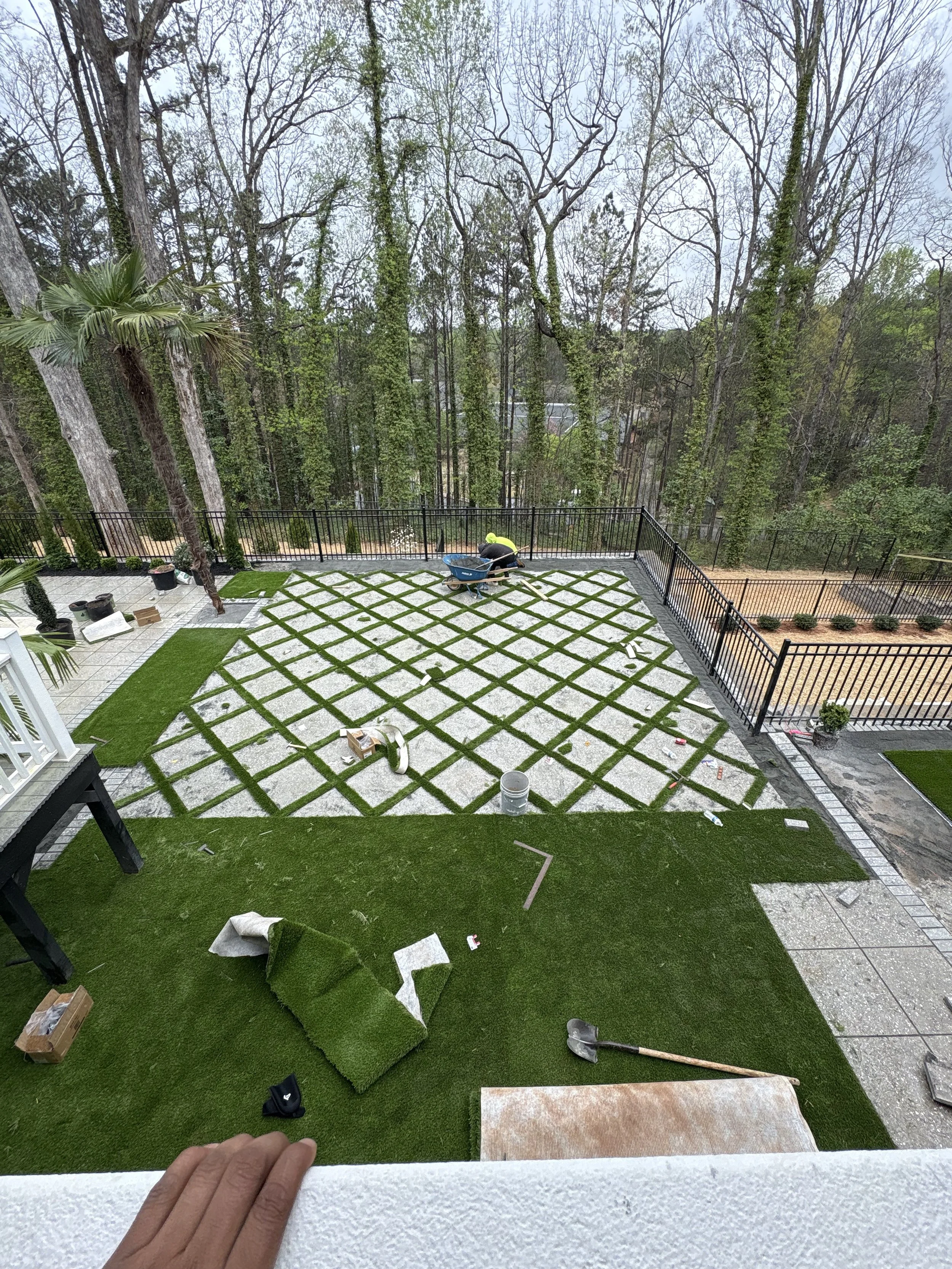 A worker installing a decorative grid pattern of grass and concrete on an outdoor patio area, with trees in the background, and tools and supplies scattered around.
