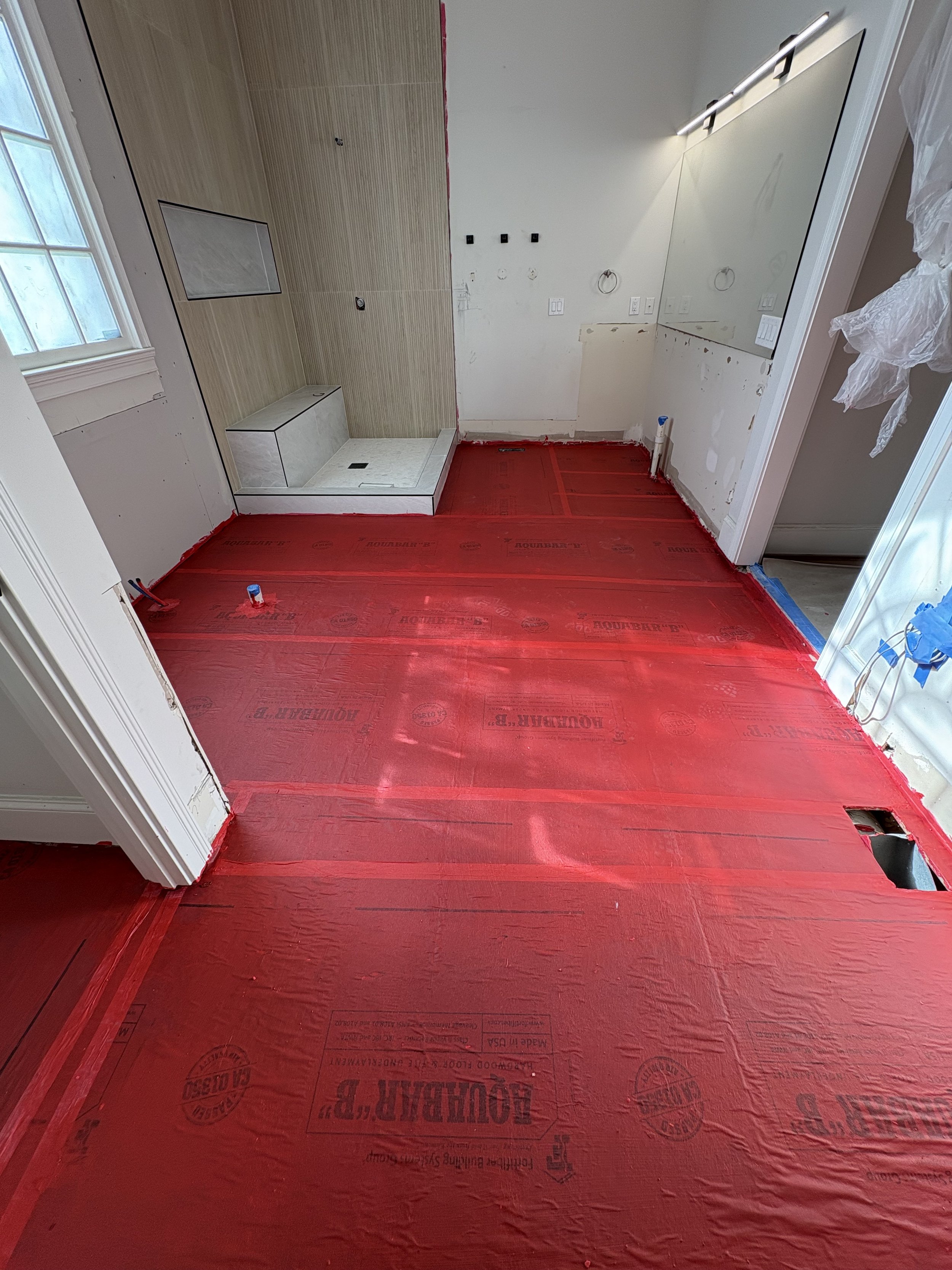 A bathroom under renovation with red vapor barrier flooring, a walk-in shower with a niche, partially installed wall tiles, and a large mirror on the wall.