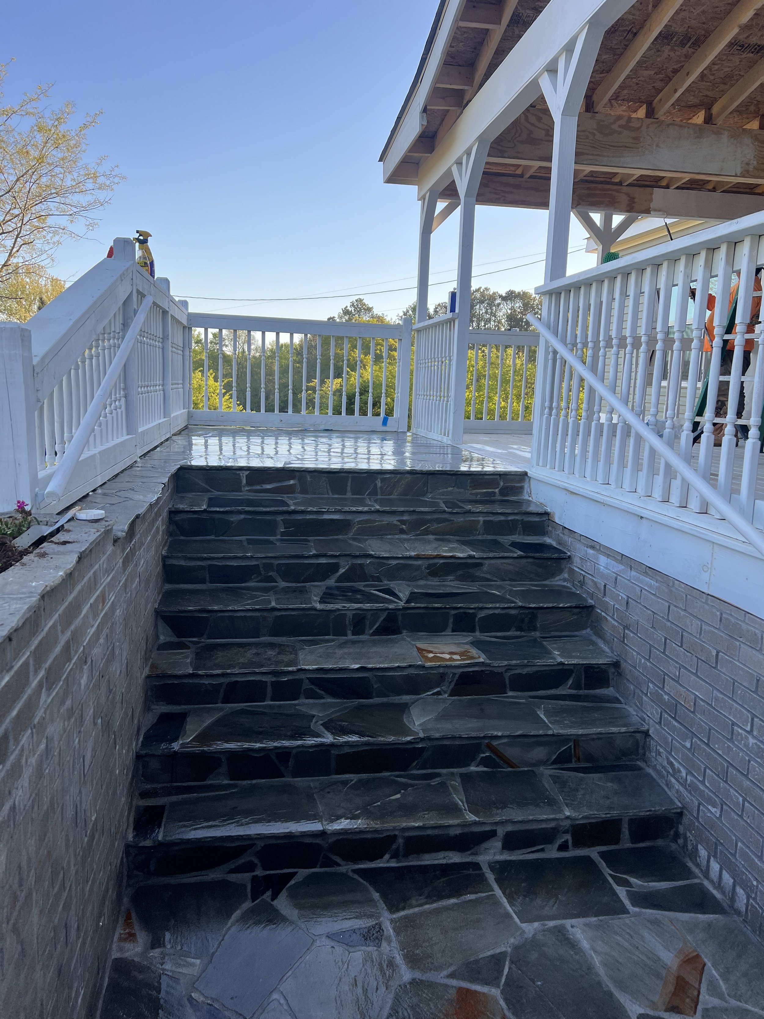 Outdoor staircase with dark stone steps leading up to a patio with white railing. Part of a house with exposed wooden under-construction porch visible on the right. Clear blue sky and green trees in the background.