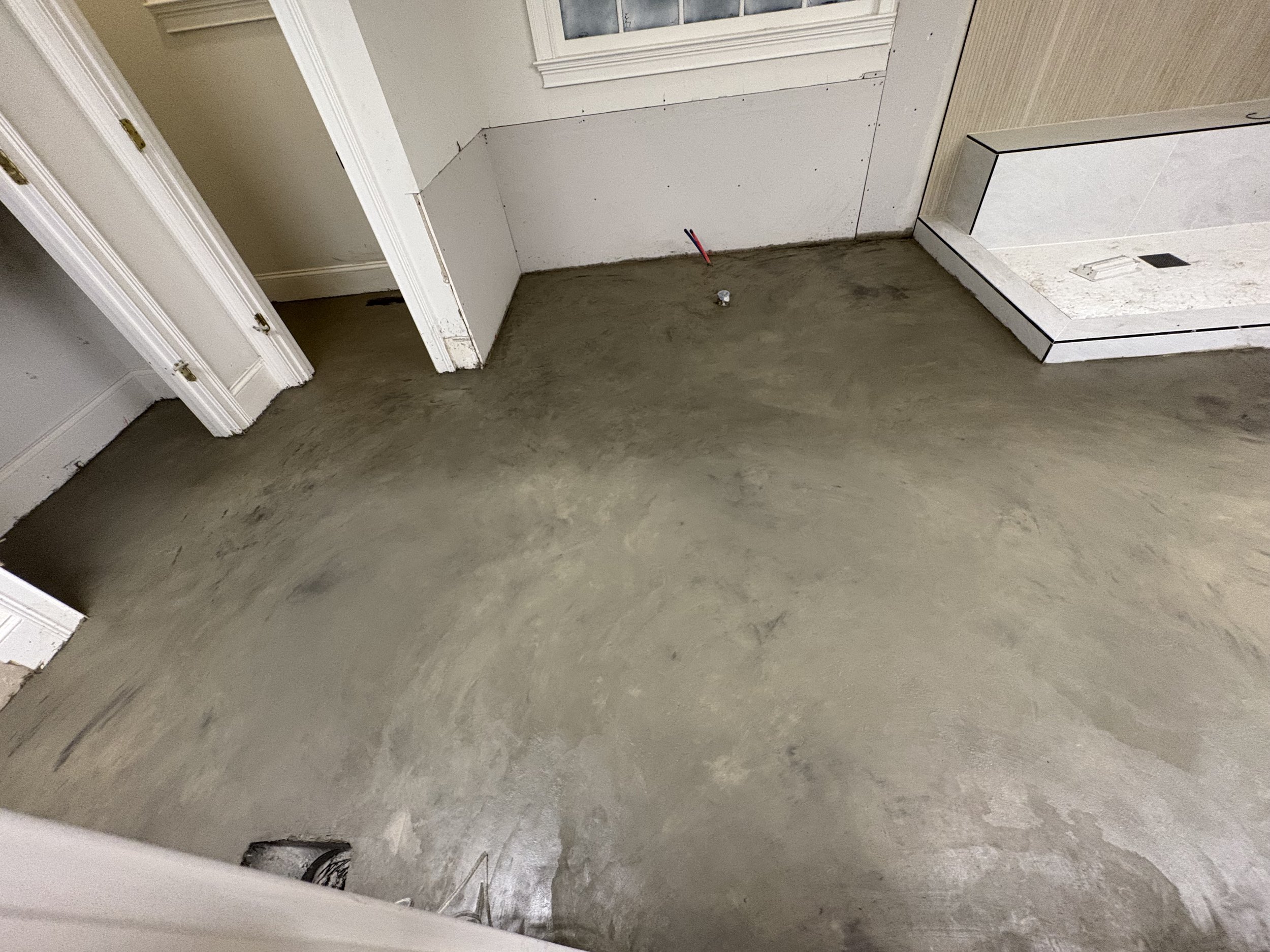 Freshly poured concrete floor in a room under construction, with walls and doorway visible, and a small section of marble tile installation in the corner.