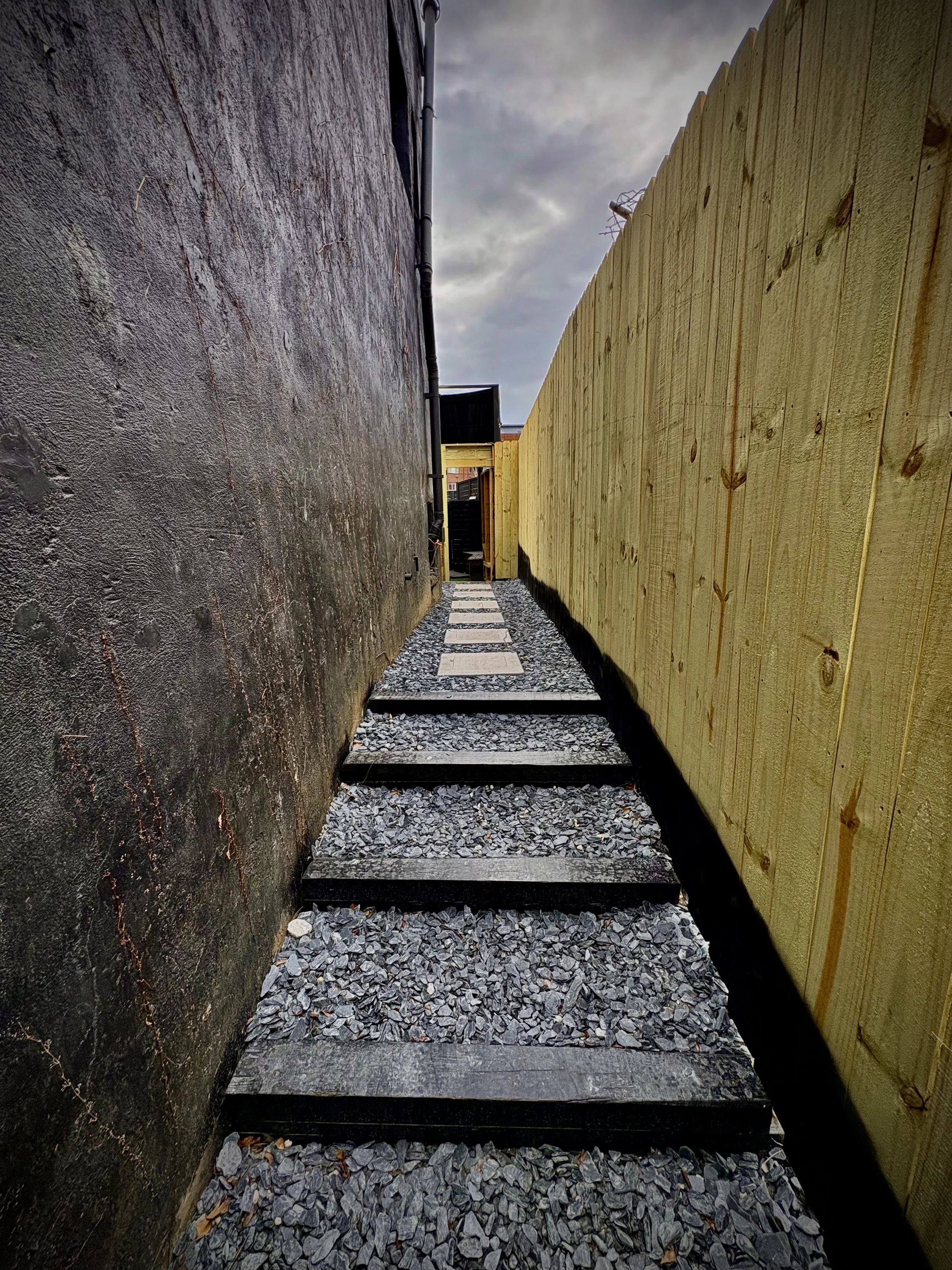 A narrow outdoor pathway with wooden stepping stones on gravel, flanked by a dark wall on the left and a light-colored wooden fence on the right, leading to a gate.
