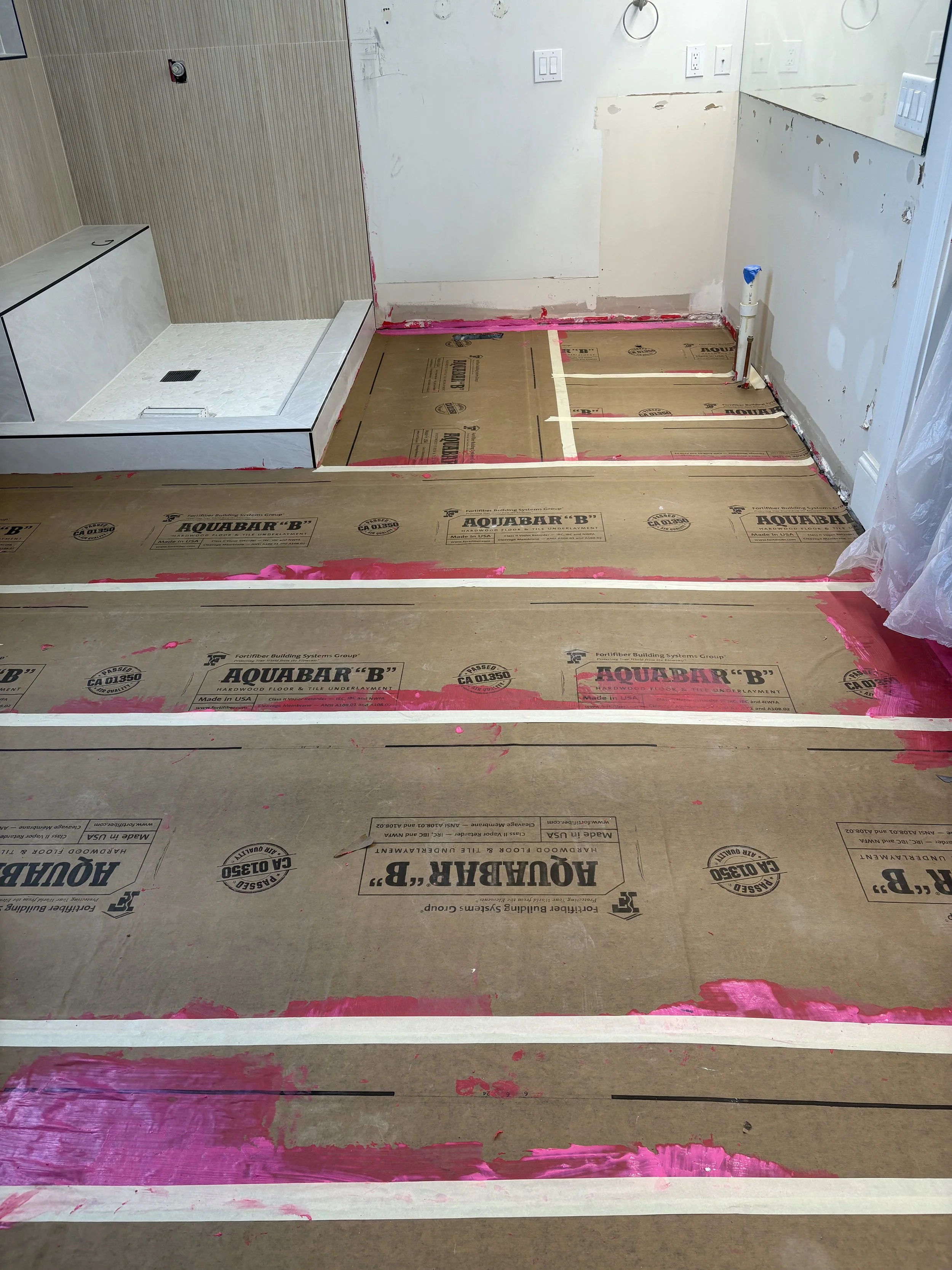 Bathroom under renovation with subflooring and pink paint patches visible, shower area in the corner, and protective plastic sheeting on side.