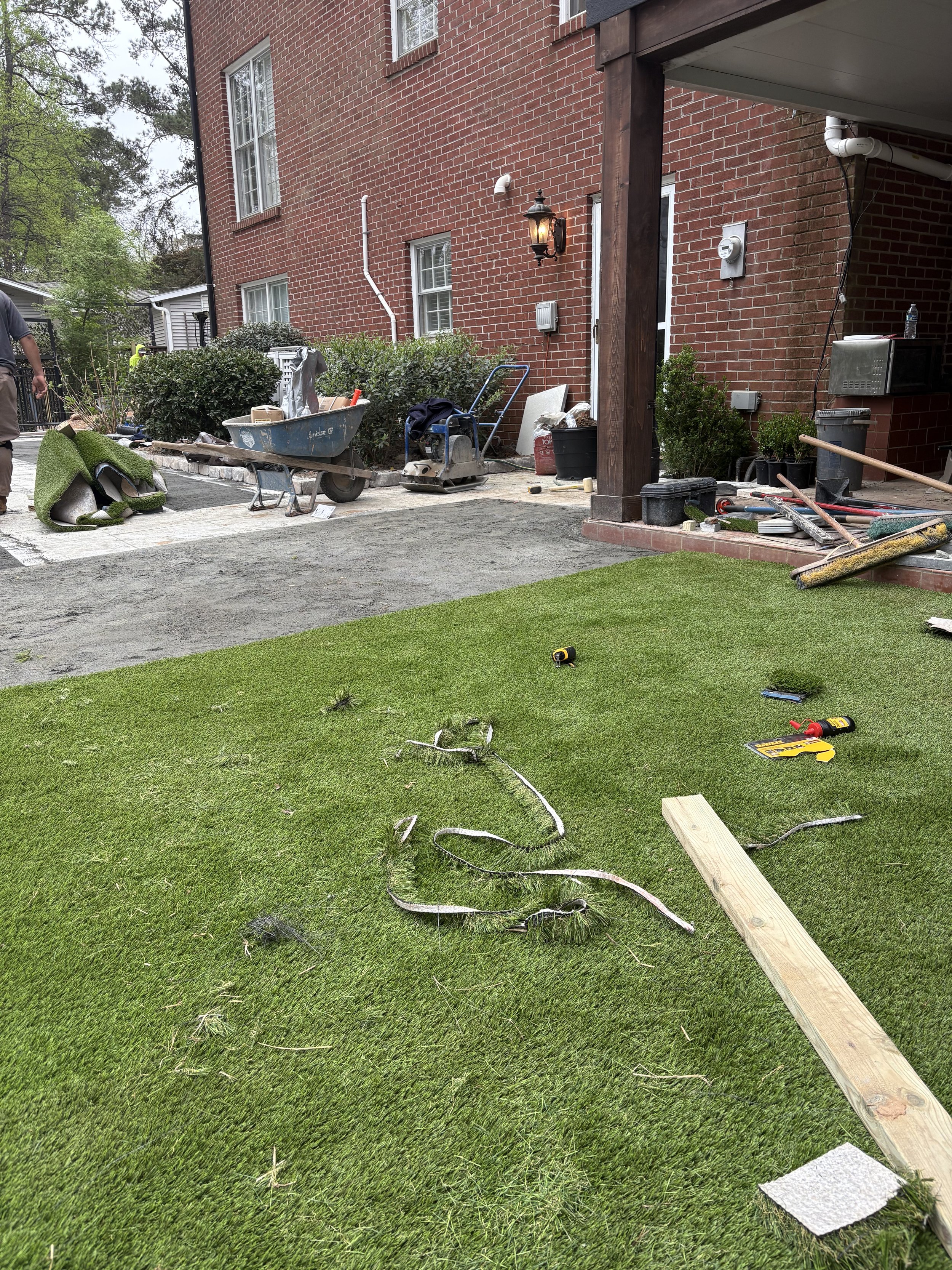 Residential backyard under construction with tools, materials, and equipment scattered on the lawn and patio, including a wooden plank, screwdrivers, and a tape measure.