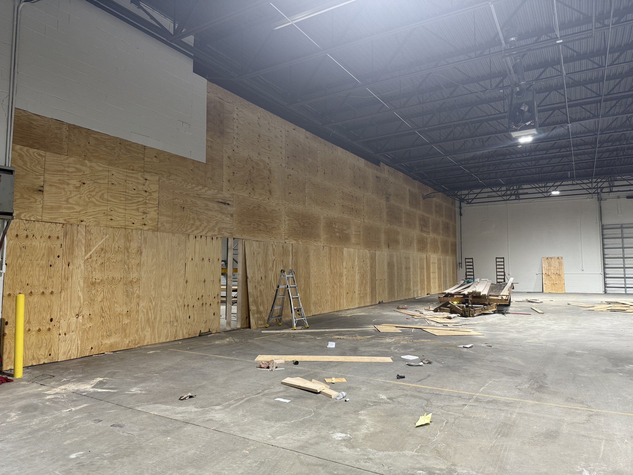 Interior of a warehouse with plywood panels being installed on the wall; construction tools and debris scattered on the concrete floor.