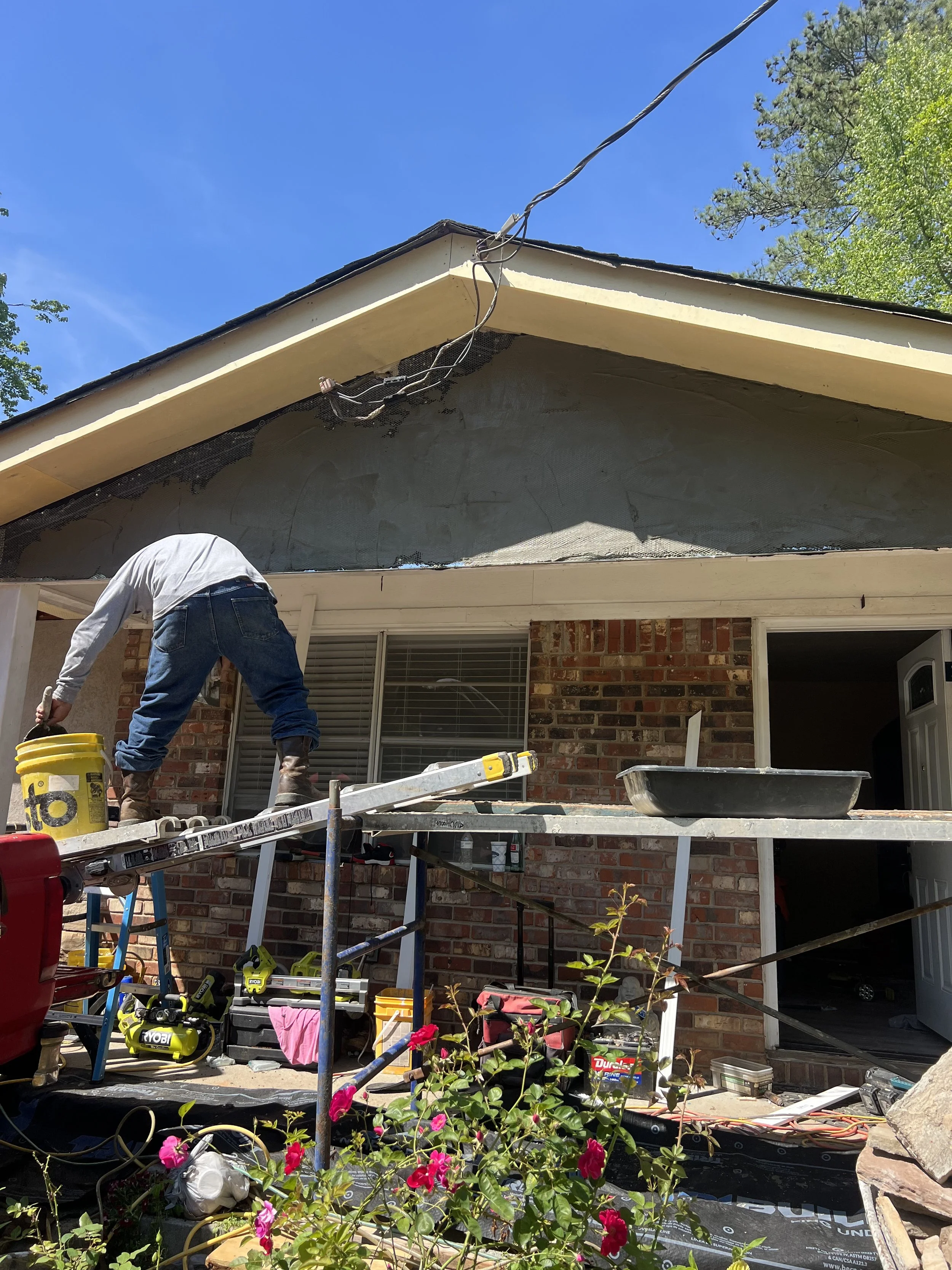 A construction worker standing on scaffolding, working on the exterior roof of a house with brick walls, with tools and supplies around.
