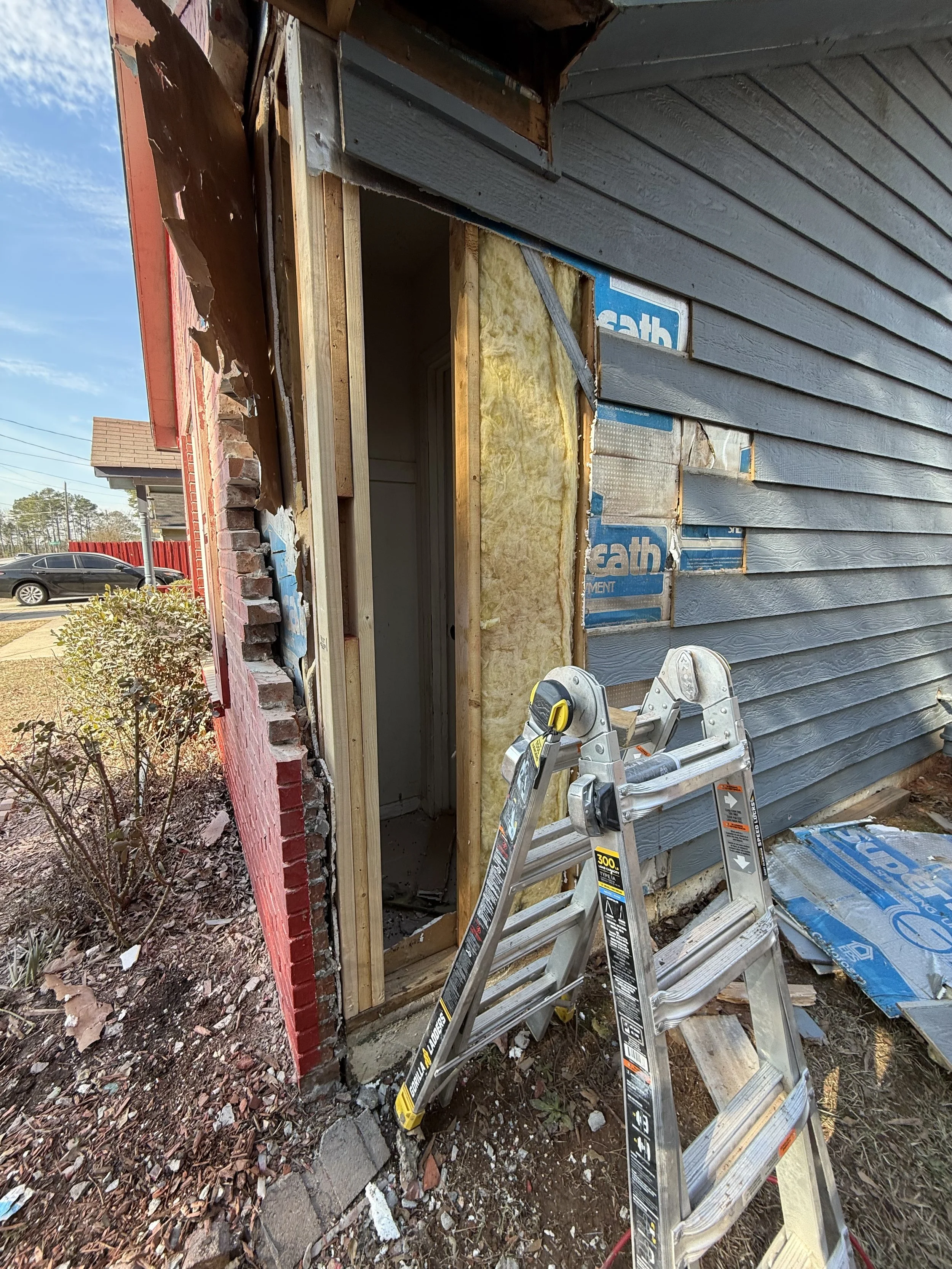Construction site showing a partially removed wall of exterior siding with insulation and inner framing visible. A ladder is placed in front, and debris is scattered on the ground. A new doorway is being built or repaired.