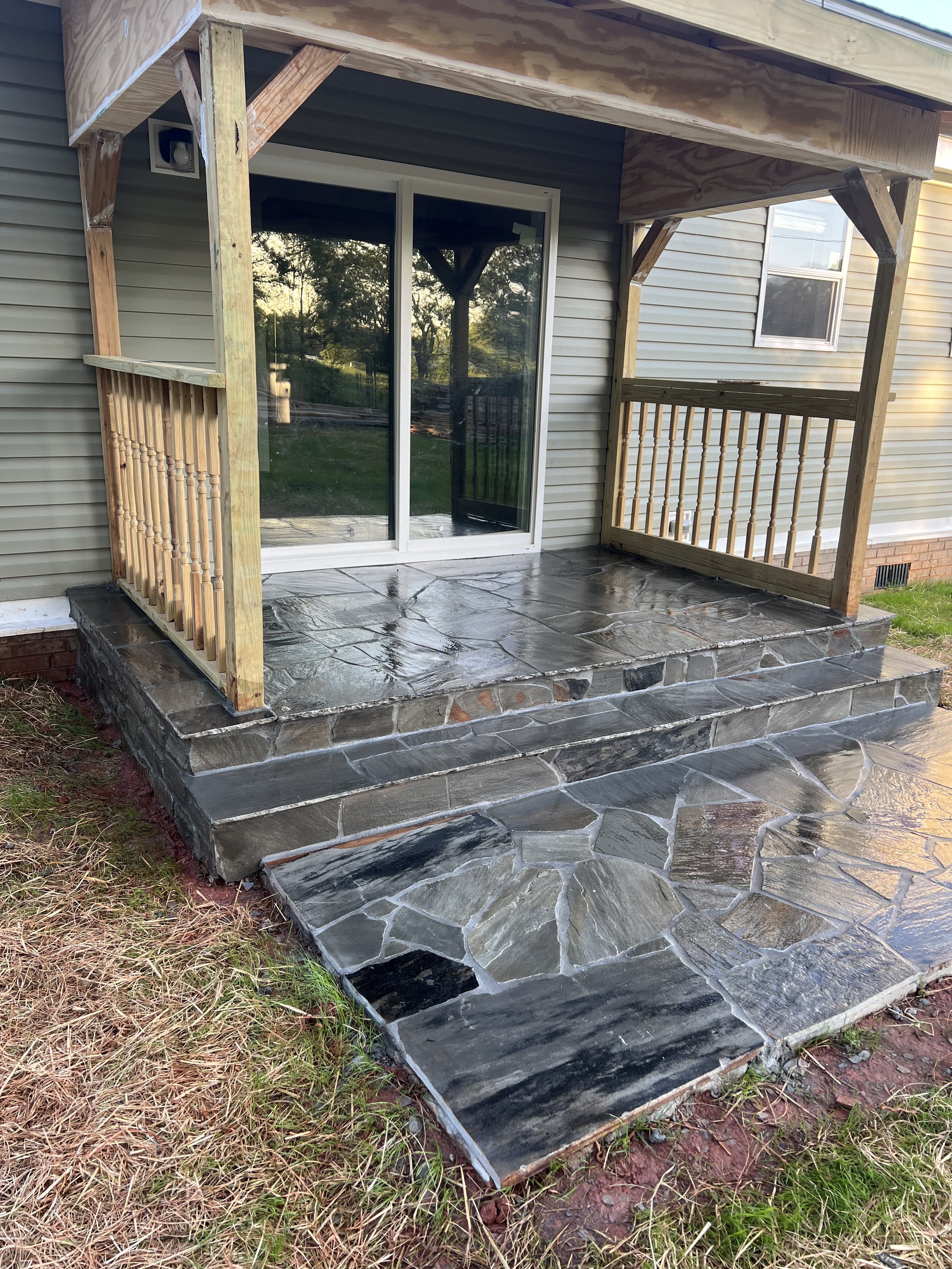 A newly constructed small porch with a stone tile floor and wooden railings, attached to a house with sliding glass doors and vinyl siding.