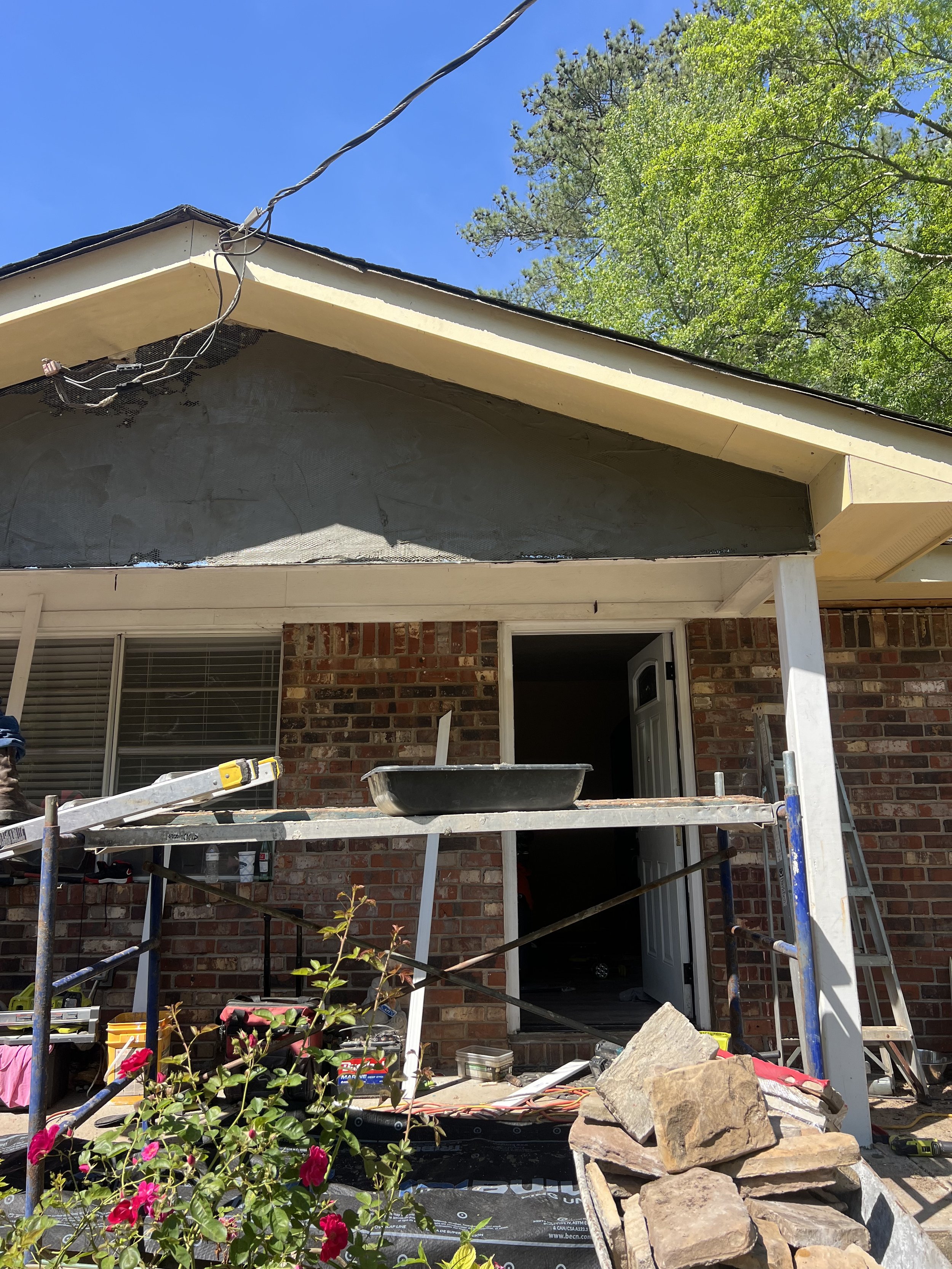 Exterior of a house under renovation with scaffolding, construction tools, and supplies visible, and pink flowers in the foreground.
