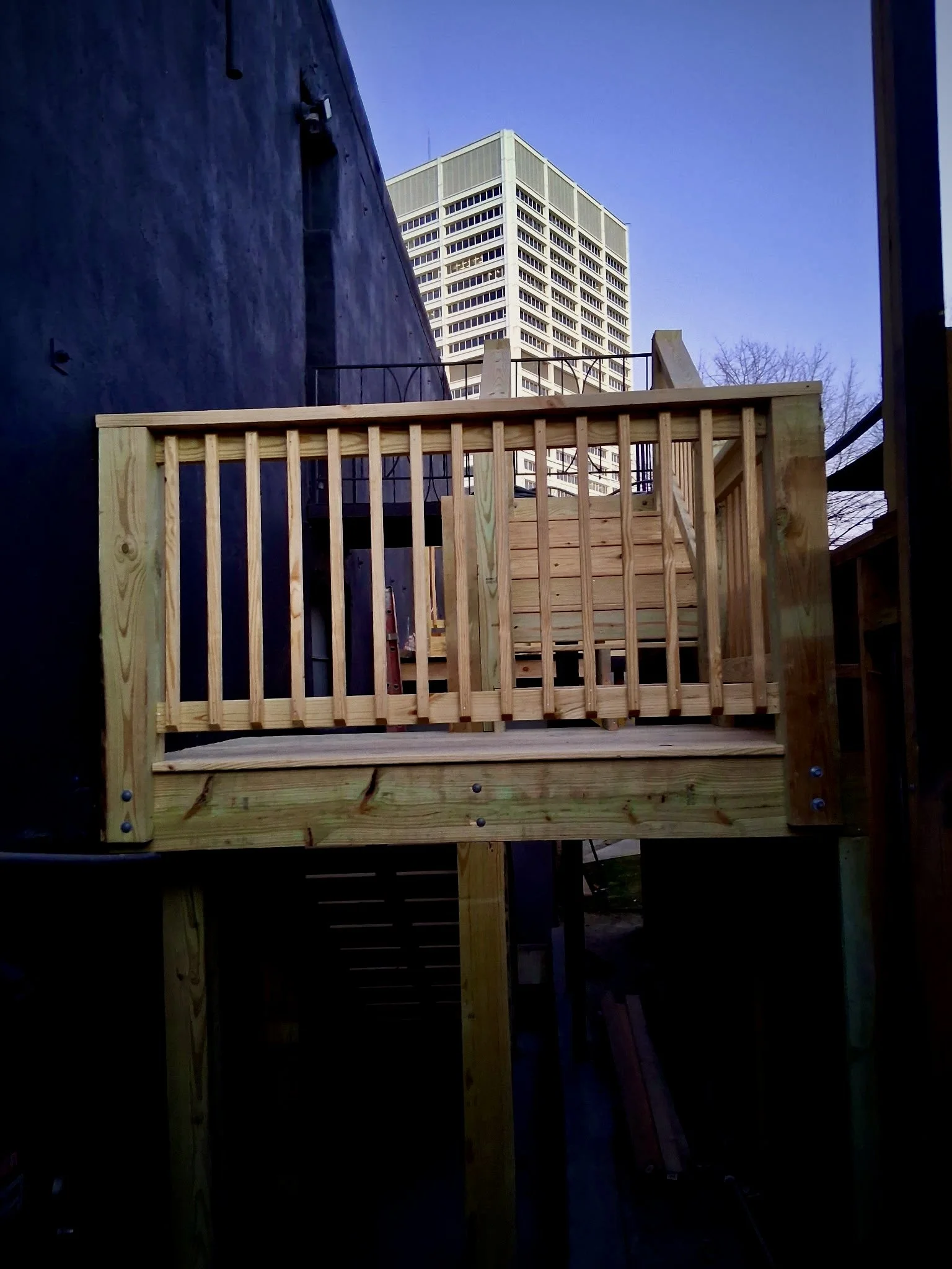 Wooden balcony with vertical railing and stairs in an urban setting, with a high-rise building in the background.