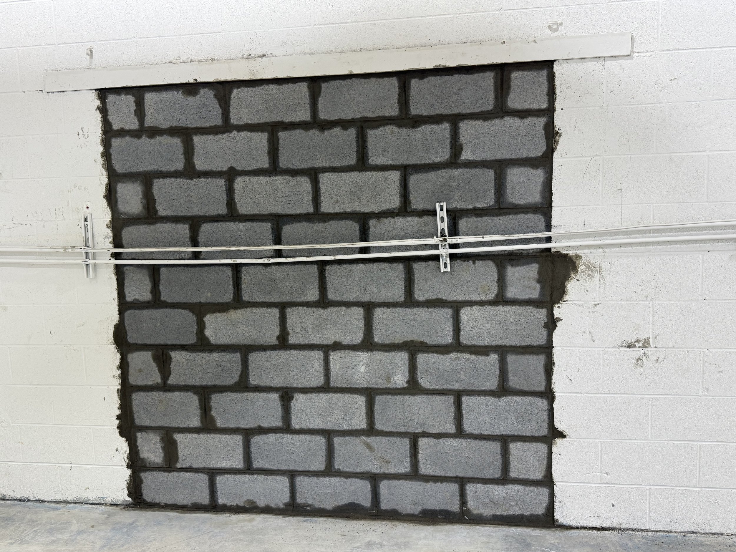 A wall with a section of gray cinder blocks, surrounded by white painted cinder blocks, with electrical conduit and a mounting bracket installed across the section.